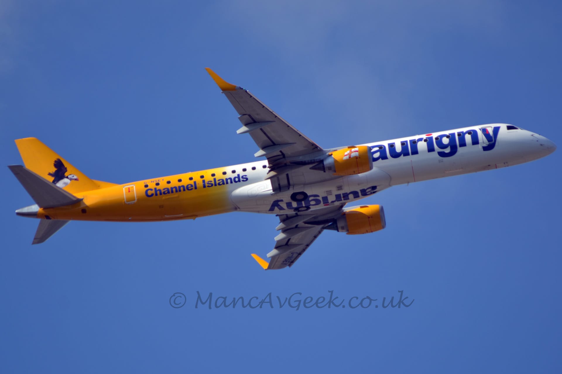 Low side view of a twin engined jet airliner flying from left to right at a very low altitude, with undercarriage stowed away, flaps extended from the rear of the wings, and the nose raised significantly, suggesting it has just taken off. The plane is mostly white, with yellow tail and engine pods under the wings. There are blue "Aurigny" titles in lower case on the forward fuselage, as well as under the wing root fairing on the belly. There is additional blue text "Channel Islands" on the lower rear fuselage, under the registration "G-NSEY". An image of a flying  black and white bird fills most of the tail. Vivid blue sky fills the rest of the frame.