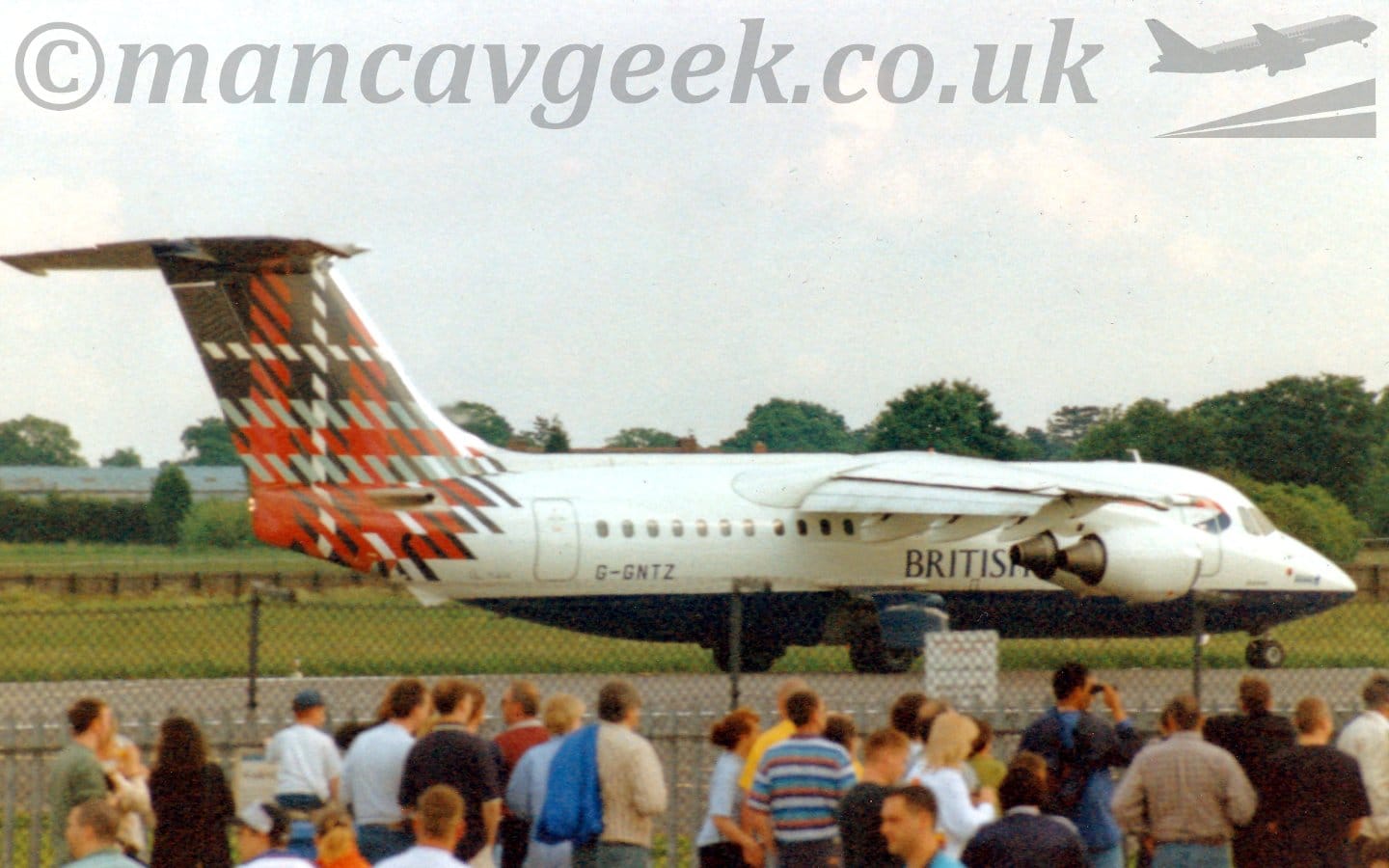 Side view of a high-winged, 4 engined jet airliner taxiing from left to right along a grey runway. The plane is mostly white, with a blue belly, and "British" titles on the lower forward fuselage. The black registration "G-GNTZ" is on the lower rear fuselage, while the tail and rear fuselage is covered in a black, grey, red, and white tartan. In the foreground, a large group of people is gathered around, some looking at this plane but mostly staring in various, different directions, with a black mesh fence between them and the runway. Green grass in the background leads up to trees in the distance, under cloudy white sky.