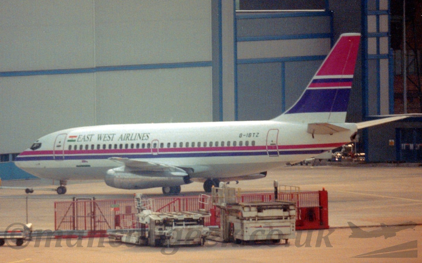 Side view of a twin engined jet airliner parked facing to the left in front of a large grey hangar. The plane is mostly white, with a pink and purple stripe running along the body, and black "East West Airlines" titles next to an Indian flag on the upper forward fuselage. The tail has more pink and purple stripes, of varying widths. The black registration "G-IBTZ" is o n the upper rear fuselage. A pair of white cargo loaders are parked in front of a red metal gate in the foreground, with a large grey hangar dominating the background.