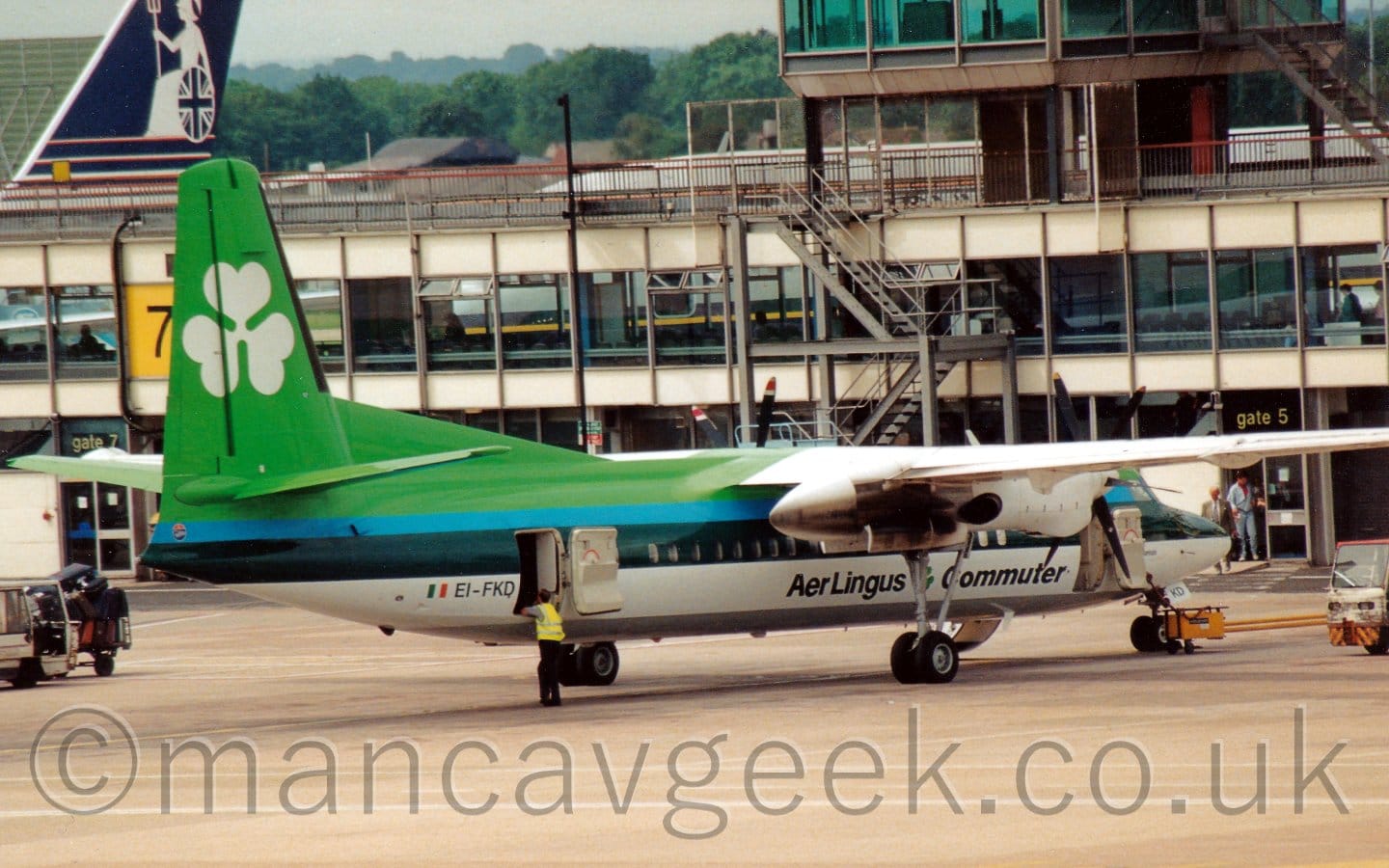 Side view of a high-winged, twin propellor-engined parked facing to the right. The plane is mostly various shades of green, with bright green at the top, darker blue-grey stripes along the middle of the body, and white at the bottom. There are black "Aer Lingus Commuter" titles on the lower forward fuselage, and a large white shamrock on the green tail. There are doors open at both the front and the rear, with a man wearing a yellow hi-viz jacket leaning on the cabin door while standing outside the rear door. Grey concrete apron fills the foreground, while a heavily-glazed terminal building stretches across the frame in the background, with the dark blue tail of a larger airliner rearing over the top of the building on the other side, with trees and grey sky in the distance.