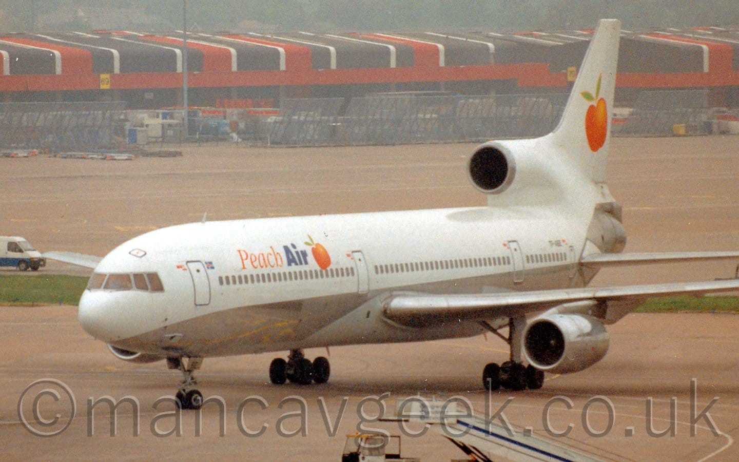 Side view of a 3 engined jet airliner taxiing from right to left. The plane is mostly white, with a silver belly. There are orange and blue "Peach Air" titles on the upper forward fuselage, next to an image of an orange coloured fruit, this logo also appearing in a larger form on the tail. Grey concrete apron fills most of the frame, with red, white, and brown cargo sheds stretching across the image in the background.