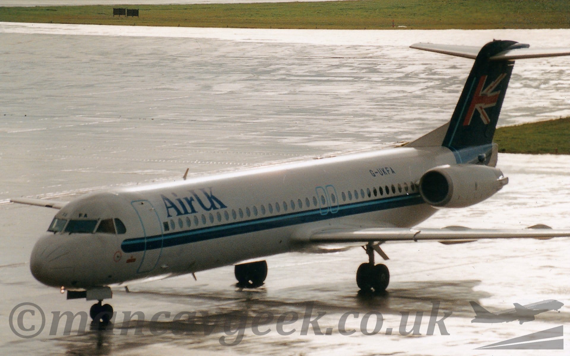 Side view of a twin engined jet airliner with the engines mounted on the sides of the rear fuselage, taxiing from right to left. The plane is mostly white, with a dark blue stripe with a pale blue pinstripe running along the body, sweeping up to fill the tail, with half a waving UK flag. There are large dark blue "AirUK" titles on the upper forward fuselage, and the black registration "G-UKFA" on the upper rear. The main door at the front and the over-wing emergency exits are outlined in a pale blue. Bright sunlight is beating down on wet concrete apron around the plane, in both the foreground and background, with large grassed areas in the background.