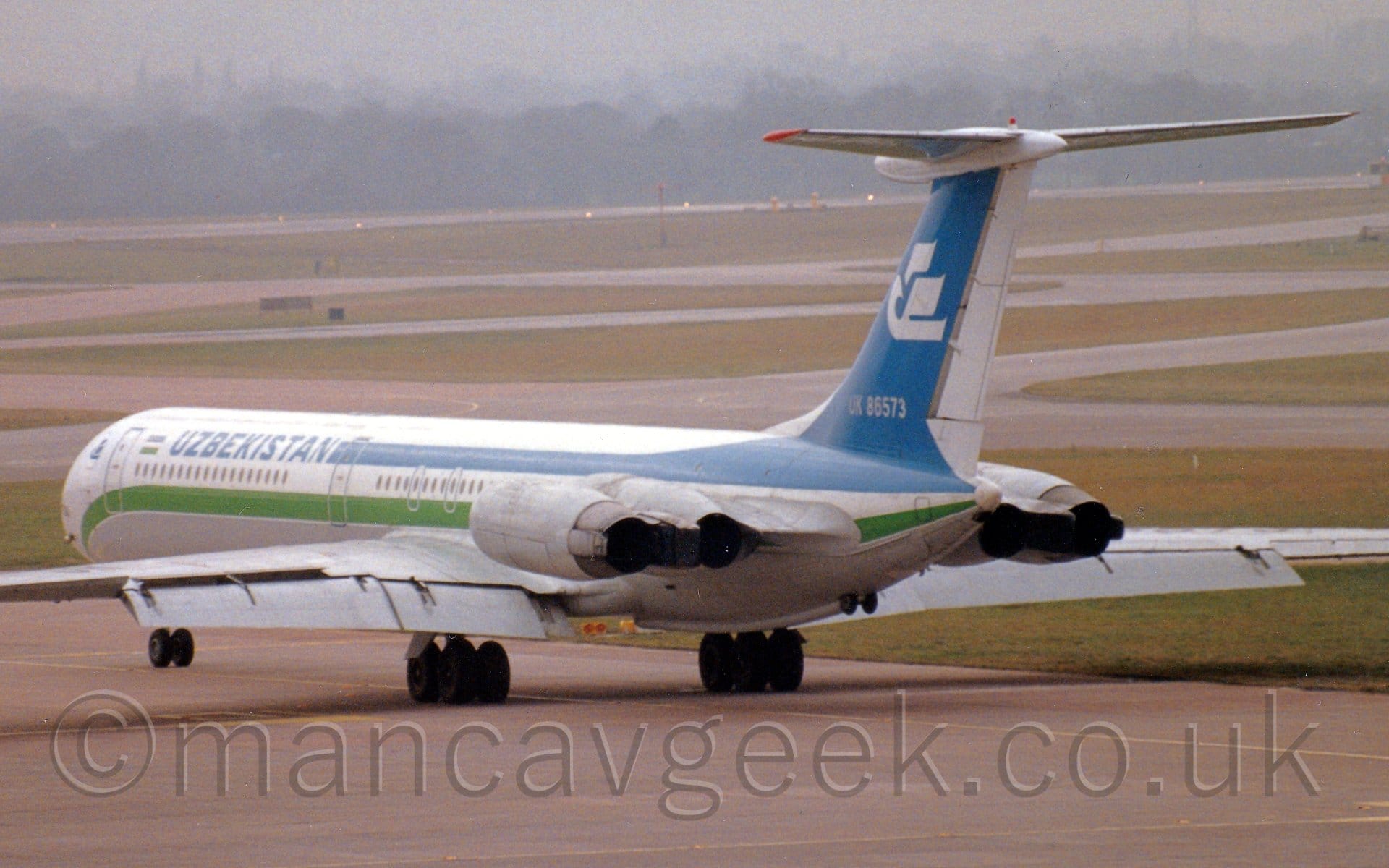 Rear side view of a 4 engined jet airliner taxiing from right to left. The plane is mostly white, with green and blue stripes running along the body, and blue "Uzbekistan" titles on the upper forward fuselage. The tail is blue, with a stylised white flying bird in the middle, and the white registration "UK-86573" at the base. The 4 engines are mounted in pairs on the sides of the rear fuselage, under the tail. Flaps are already extended from the rear of the wings, even though it has a long way to go before it gets to the runway. Grey concrete apron fills the foreground, while taxiways wend their way through large grassed areas in the background, leading up to trees vanishing into haze in the distance, under grim grey sky.