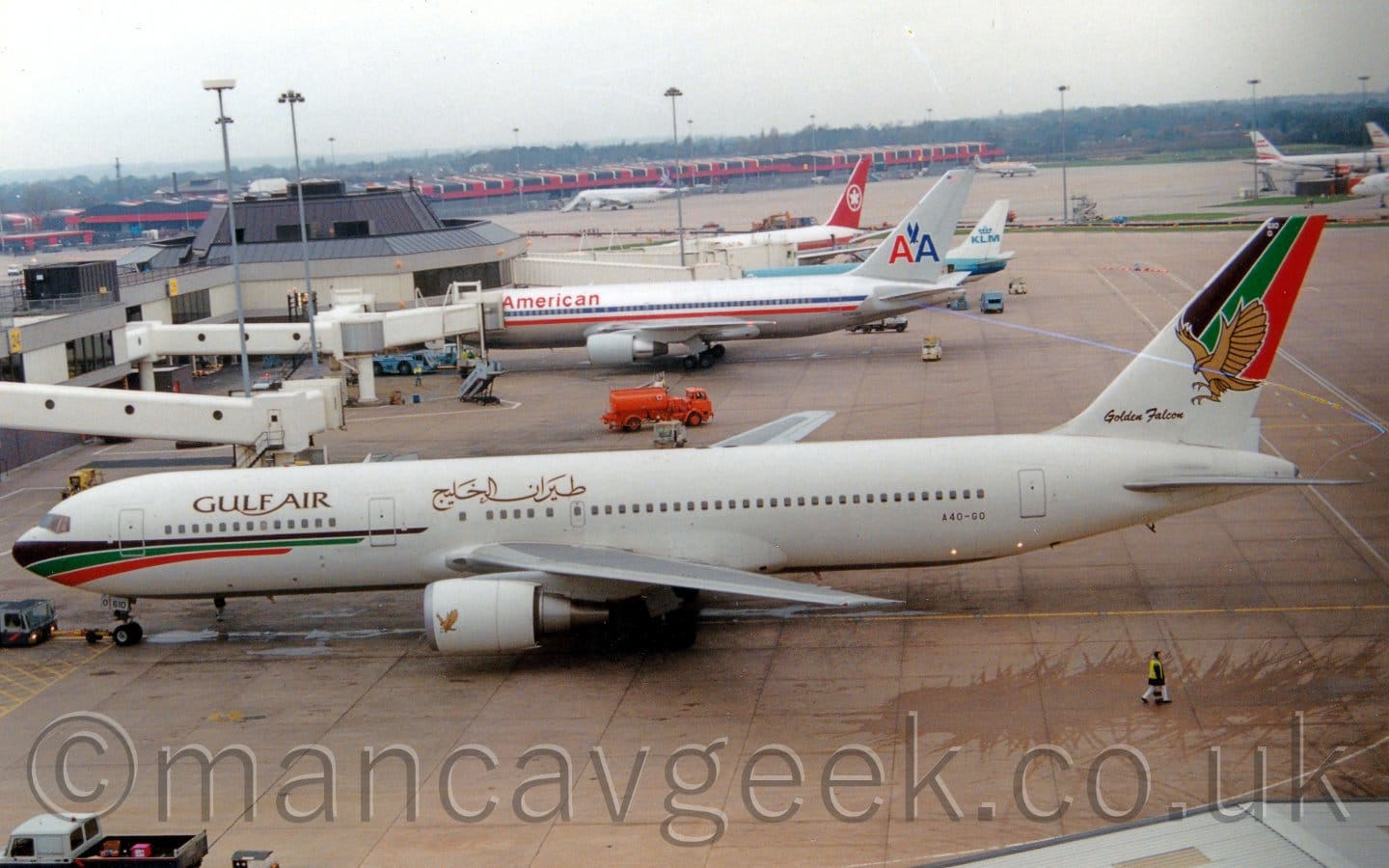 Side view of a twin engined jet airliner facing to the left but being pushed back to the right by a low-slung, grey and dark blue tag attached to the nose-wheel by a long yellow pole. The plane is mostly white, with gold "Gulf Air" titles on the upper forward fuselage, in English at the front, and in an Arabic script over the wing. There are black, green, and red stripes wrapped around the nose, as well as covering the top of the tail. A large golden image of a stooping falcon fills most of the tail, with "Golden Falcon" text, also in gold, at the base of the tail. A smaller golden falcon is at the front of the white engine pods. Grey concrete apron fills most of the frame, with a white and grey terminal building stretching into the distance, with a handful of jet airliners parked facing it. Some more airliners are parked in the distance, including by the red, black, and white cargo sheds in the distance. Bright but hazy sky fills the rest of the frame.
