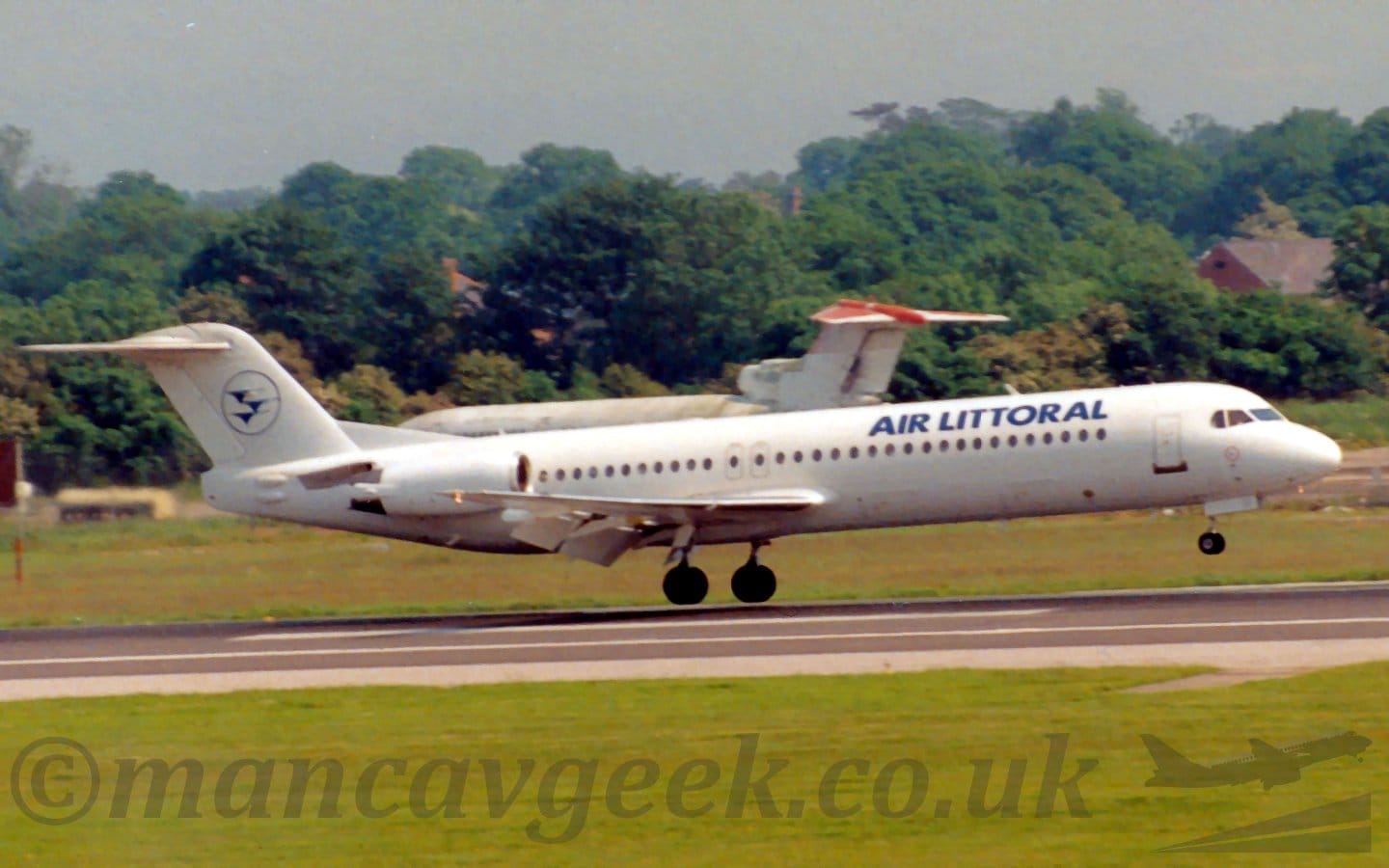 Side view of a white, twin engined jet airliner with the engines mounted on the sides of the rear fuselage, flying from left to right at a very low altitude, maybe a metre above as runway, with the undercarriage lowered, flaps extended from the rear of the wings, and the nose fairly flat, suggesting it is about to land. Green grass fills the foreground, lining the side of the runway, with more grass in the background, leading up to trees in the background. The rather dilapidated white fuselage of a 3 engined jet airliner is visible in front of the trees, facing to the left. Grey sky fills the rest of the frame.