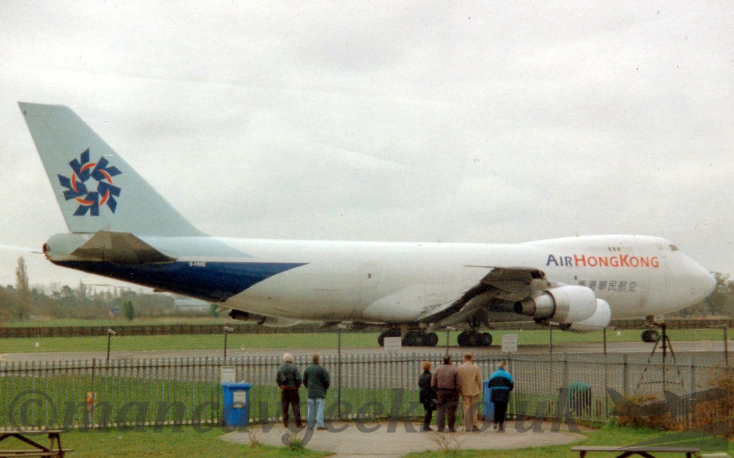 Side view of a very large, 4 engined jet airliner taxiing from left to right. The plane is mostly white, with blue and red "Air Hong Kong" titles on the upper forward fuselage, with some writing in a blue Chinese script below. The planes tail is a pale blue, with dark blue arrow heads arranged in a circle, , pointing inwards, with red lines connecting them. The lower rear fuselage is a dark blue, with the white registration "B-HME" at the top. In the foreground, a group of people are standing by a tall black metal fence at the far end of the beer garden, looking at the plane, bundled up in big coats, which could mean it's winter, or it could just mean its a Tuesday in Manchester. Grey sky fills the rest of the frame.