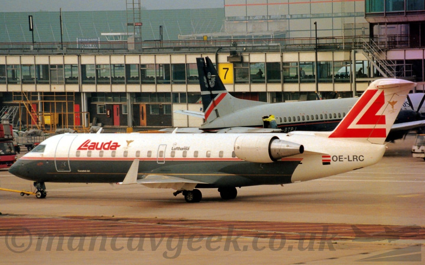 Side view of a twin engined jet airliner with the engines mounted on the sides of the rear fuselage, facing to the left but being pushed back to the right by an out-of-frame tug attached to the nose-wheel by a long yellow pole. Grey concrete apron fills the foreground, with a red strip in the middle with yellow hatching. A grey and white terminal building with heavily glazed sections stretches across the frame, with a dark blue and grey airliner on the right of the frame facing the building. A large grey hangar on the right in the distance is balanced by a large, green, boxy metal frame on the left, under grey-blue sky.