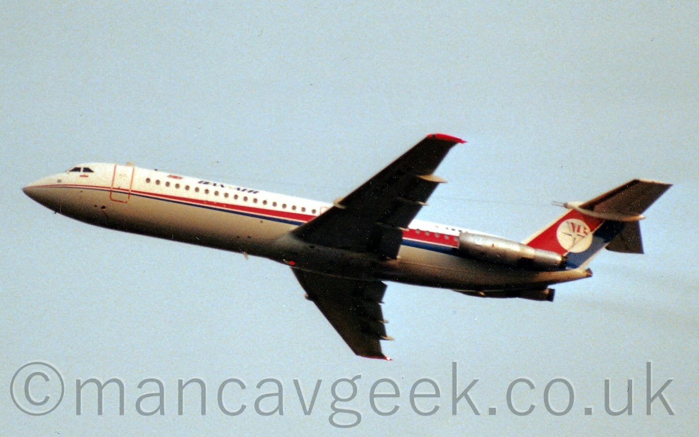 Side view of a twin engined jet airliner flying from right to left at a very low altitude, with flaps extended from the rear of the wings, undercarriage already retracted, and nose raised sharply, suggesting it has just taken off. The plane is mostly white, with a a grey belly, and a thin red and blue stripe running along the body, getting wider as it moves rearwards, until it sweeps up to fill the tail. There are blue "Dan Air" titles on the upper forward fuselage, and a white circle containing a blue 4-pointed star with a red and white striped flag on the tail. The engines mounted on the sides of the rear fuselage are in a bare metal scheme, and appear to have had rear extensions fitted with hush-kits to keep the noise from these venerable engines as low as possible. Pale blue sky fills the rest of the frame.