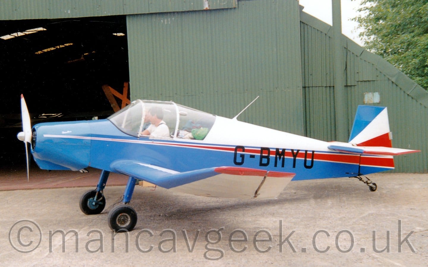 Side view of a single-engined light aircraft parked facing to the left on a black tarmac apron. The plane has a white upper rear fuselage, with most of the rest being blue, with an additional red stripe running along the body and getting wider as it moves rearwards. The black registration "G-BMYU" is overlaid on the blue and red sections on the rear fuselage. 2 people can be seen in the cockpit, with a load of bags visible in the rear of the cabin. Grey concrete apron fills the foreground, with a green arched hangar with its doors slid wide open in the background, under bright but hazy skies.