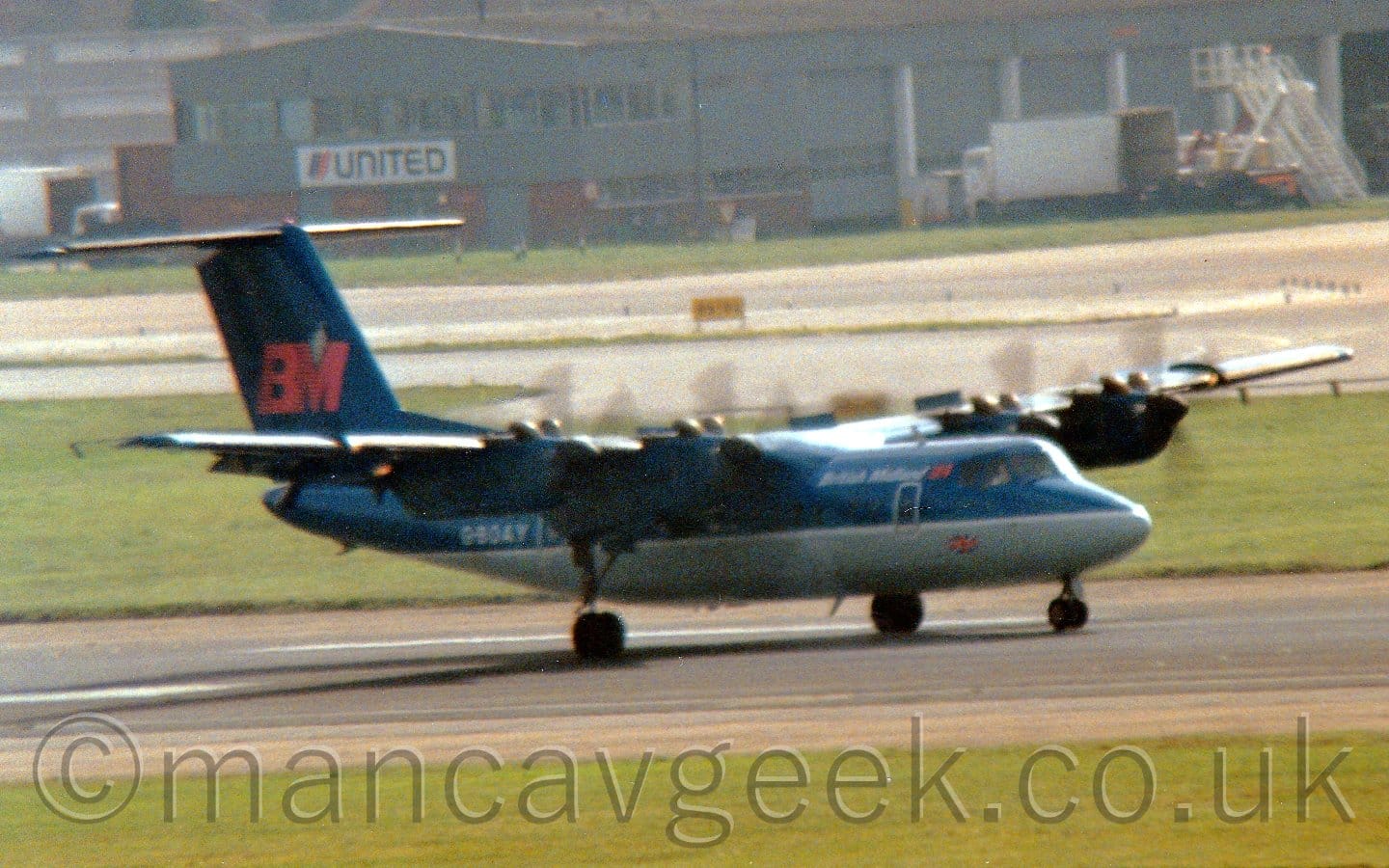 Side view of a high-winged, 4 propellor-engined airliner moving at speed from left to right along a black runway, with the propellors merely spinning blurs, and flaps extended from the rear of the wings. The plane is mostly dark blue, with a grey belly, with white "British Midland" titles on the upper forward fuselage, next to a red "BM" logo just behind the cockpit windows. The white registration "G-BOAY" is on the rear fuselage, while the tail has a larger version of the red "BM" logo. Green grass lines the sides of the runway in the foreground, with a taxiway and a grey building in the background.