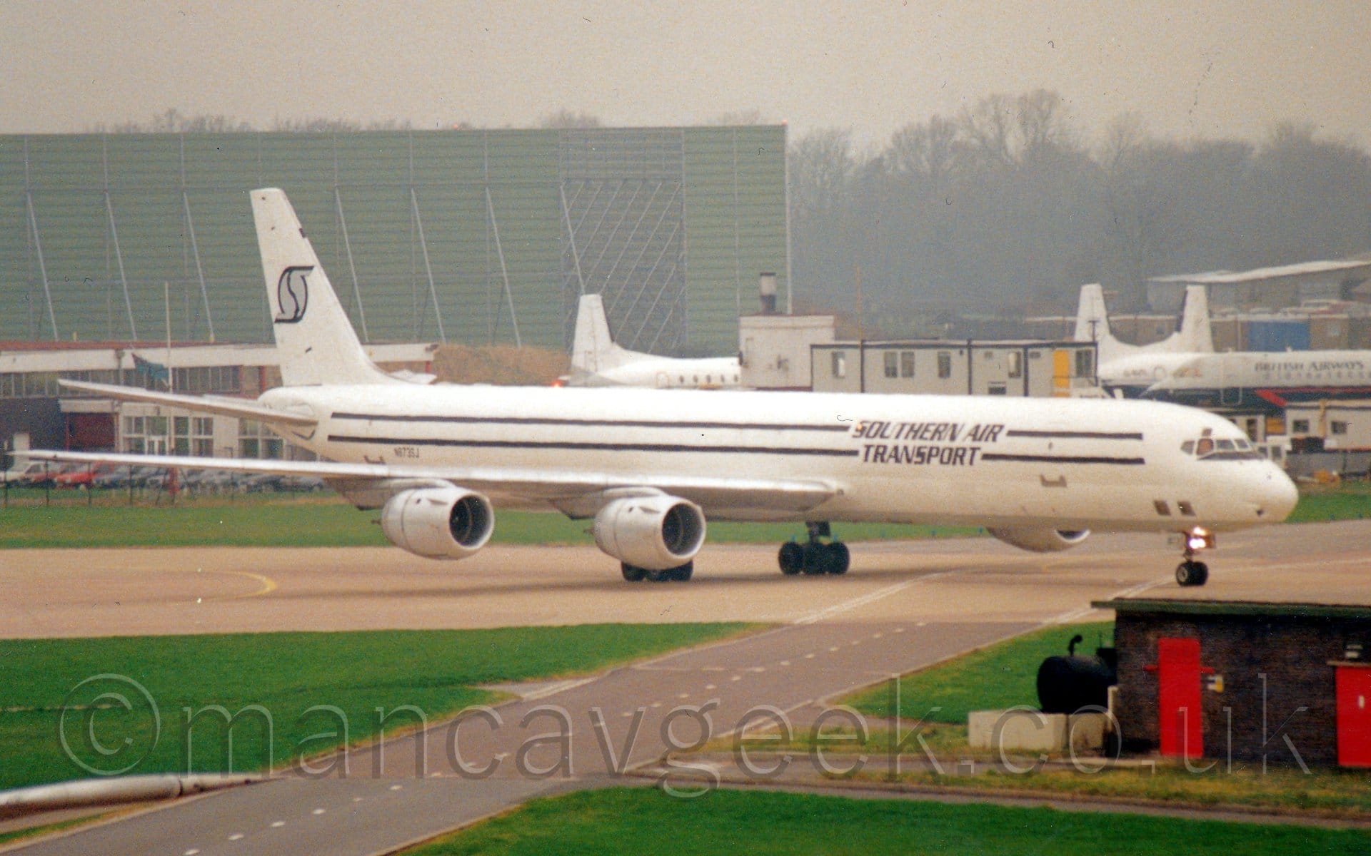 Side view  of a 4 engined jet cargo plane taxiing from left to right. The plane is mostly white, with a grey belly, and a pair of black stripes running along the body, overlaid with black "Southern Air Transport" text. The registration "N873SJ" is on the lower rear fuselage, with a stylised, fat, grey letter "S" outlined in black. Green grass fills most of the foreground, with a grey roadway cutting through, next to a low, black brick building on the right of the frame. In the background, a collection of white or grey and blue airliners are parked on the right, with a green, metal, box-like structure towering over everything on the left, with trees and grey sky filling the rest of the frame.