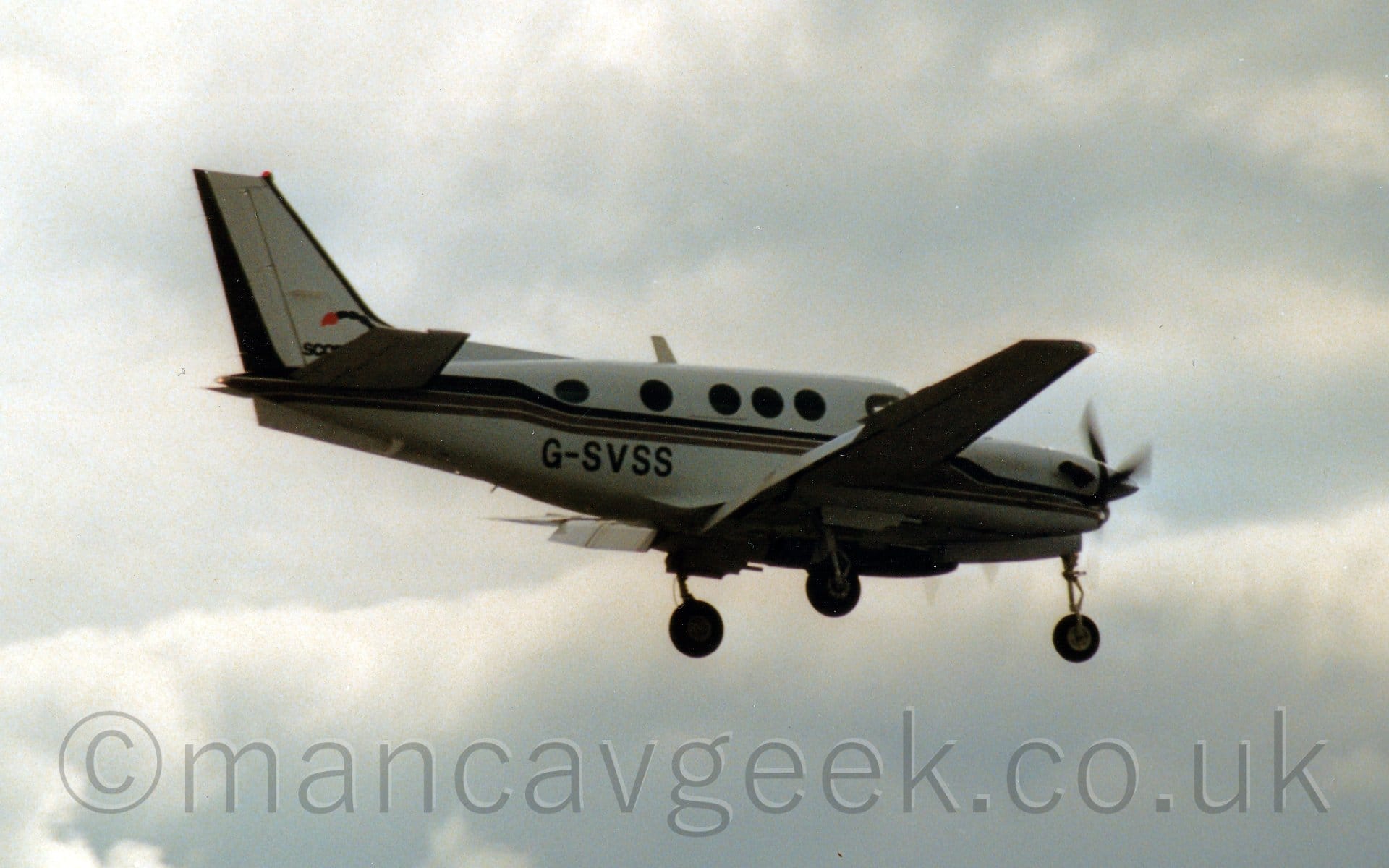 Side view of a twin propellor-engined BizProp flying from left to right at a low altitude, with flaps extended from behind the wings, and the under-carriage lowered, suggesting it is about to land. The plane is mostly white, with a dark stripe running along the body, as well as on the engine pods, with the black registration "G-SVSS" on the lower rear fuselage. Cloudy grey skies fill the rest of the frame.