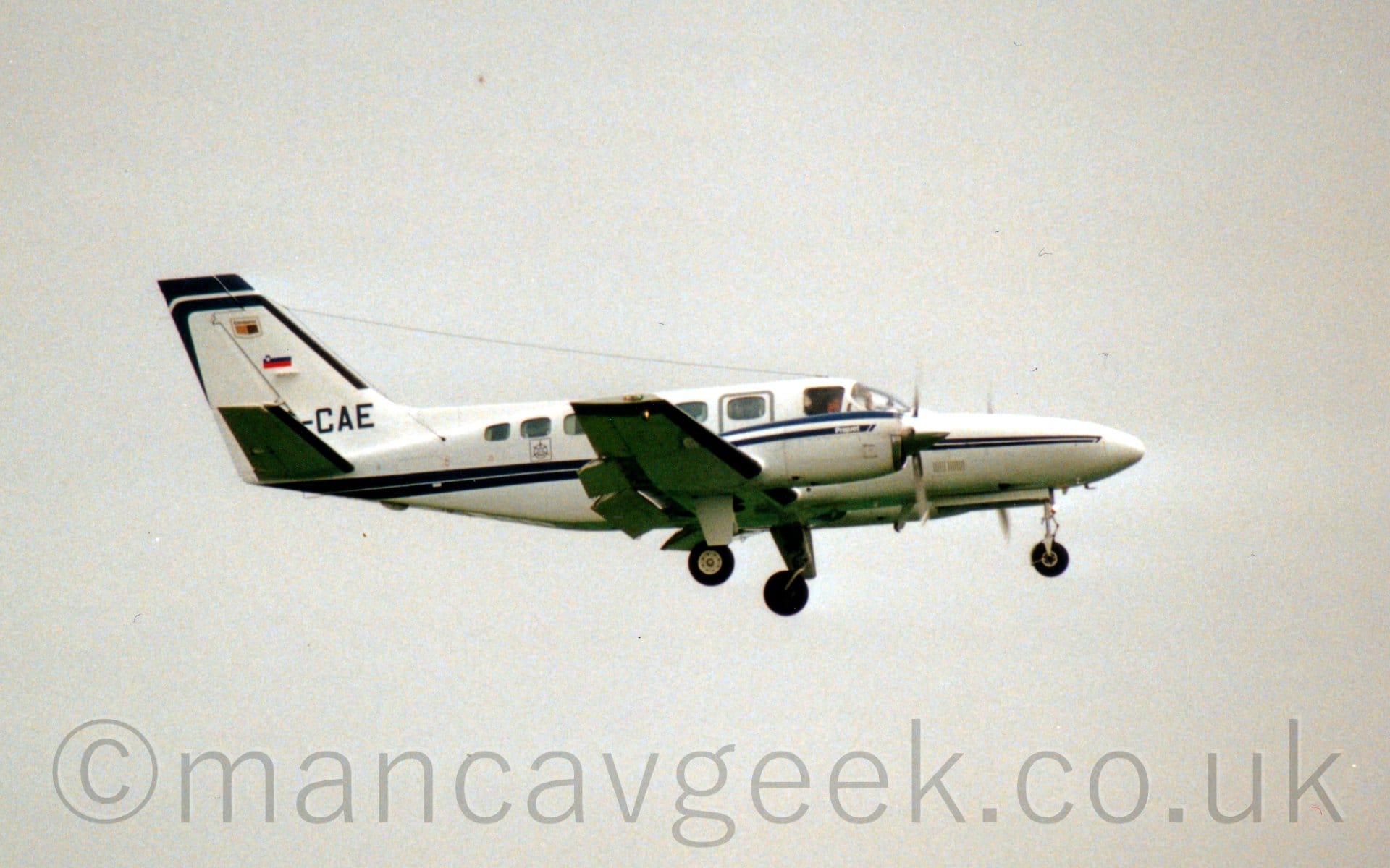 Side view of a twin engined BizProp flying from left to right at a low altitude with undercarriage lowered and flaps extended from the rear of the wings, suggesting it is about to land. The plane is mostly white, with blue stripes running along the body and over the engines, with the last 3 letters of the registration "-CAE" visible at the bottom of the tail, under a small Slovenian flag. Flat white sky fills the rest of the frame.