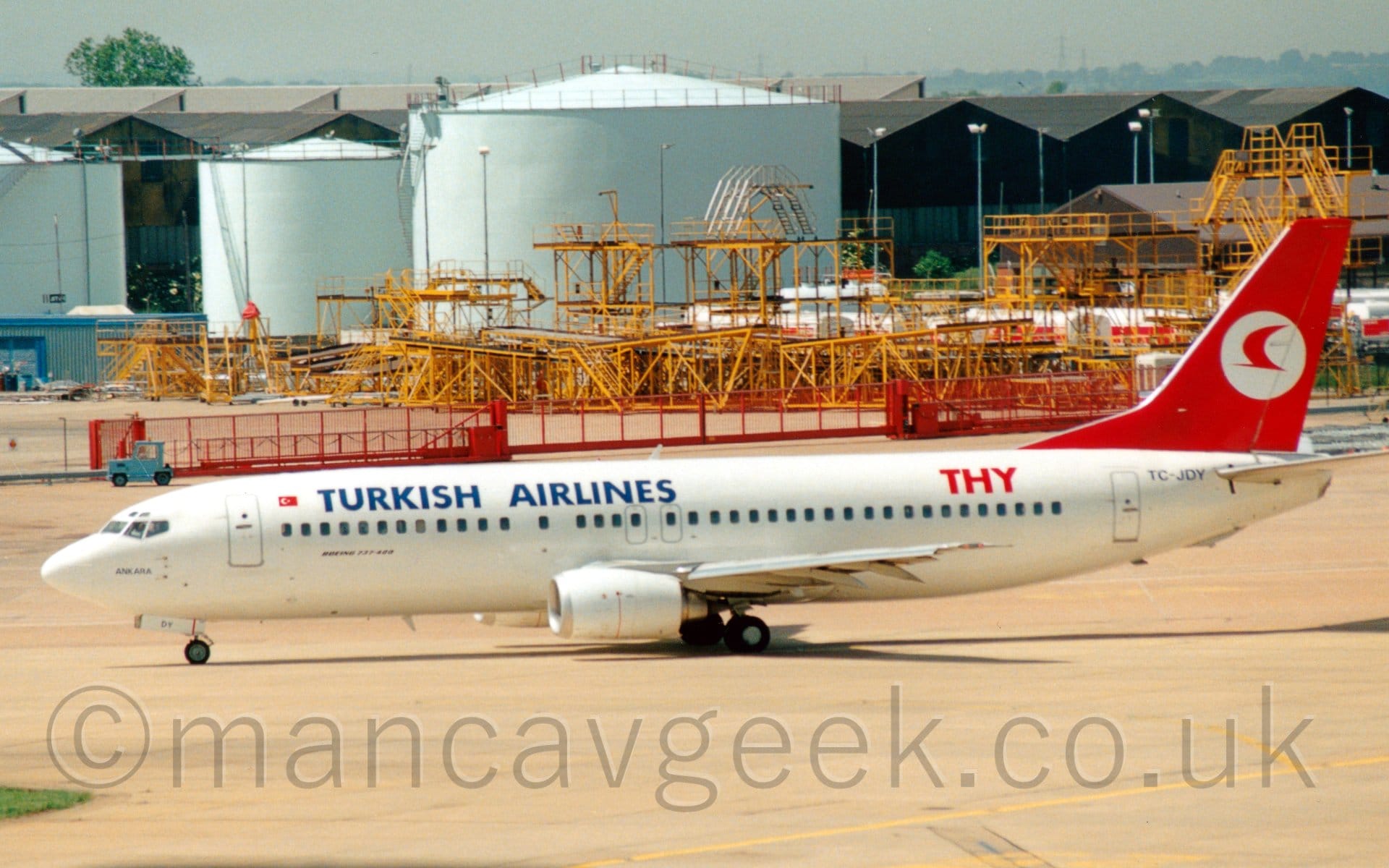 Side view of a twin engined jet airliner taxiing from right to left. The plane is mostly white, with large blue "TurkishAirlines" titles on the upper forward fuselage, red letters "THY" on the upper rear, and the black registration "TC-JDY" under the tail.. The tail is red, with a stylised red flying bird in a white circle. Grey concrete apron fills the foreground, with a collection of bizarre-looking yellow metal frames in the background, in front of so duck-egg blue fuel tanks. Black hangars stretch across the frame beyond that, under typically grey Mancunian sky.