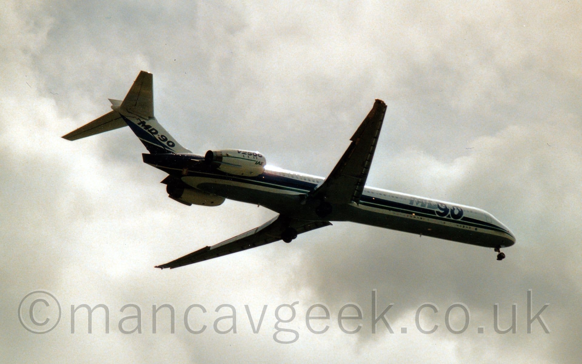 Low side view of a twin engined jet airliner flying from left to right at a very low altitude, with flaps extended from the front and rear of the wings, and undercarriage lowered, suggesting it is about to land. The plane is mostly white, with a pair of blue stripes running along the body and a grey belly. There are large, grey and blue "MD90" titles on the forward fuselage, with similar blue titles on the tail. There is blue text "V2500" on the white engine pods mounted on the sides of the rear fuselage, with smaller green "IAE" titles at the front of the engine. Somewhat dramatic grey sky fills the rest of the frame.