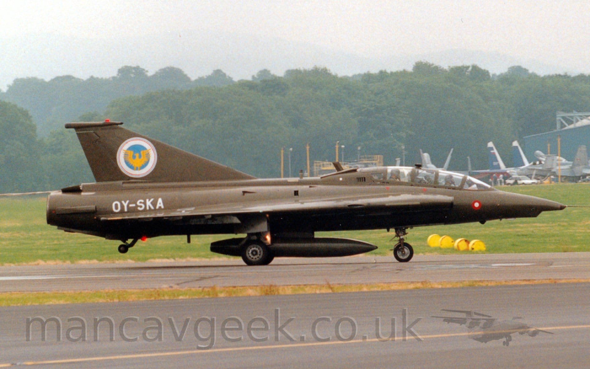 Side view of a military single engined jet fighter taxiing from left to right along a black runway. The plane is in an olive green camouflage, with the white registration "OY-SKA" on the side of the rear fuselage, and a white, blue, and yellow crest on the tail. The plane has a tandem 2 seat cockpit, one pilot sitting behind and above the other. A black taxiway fills the foreground, with grass in the background leading up to trees in the distance, under grey sky.