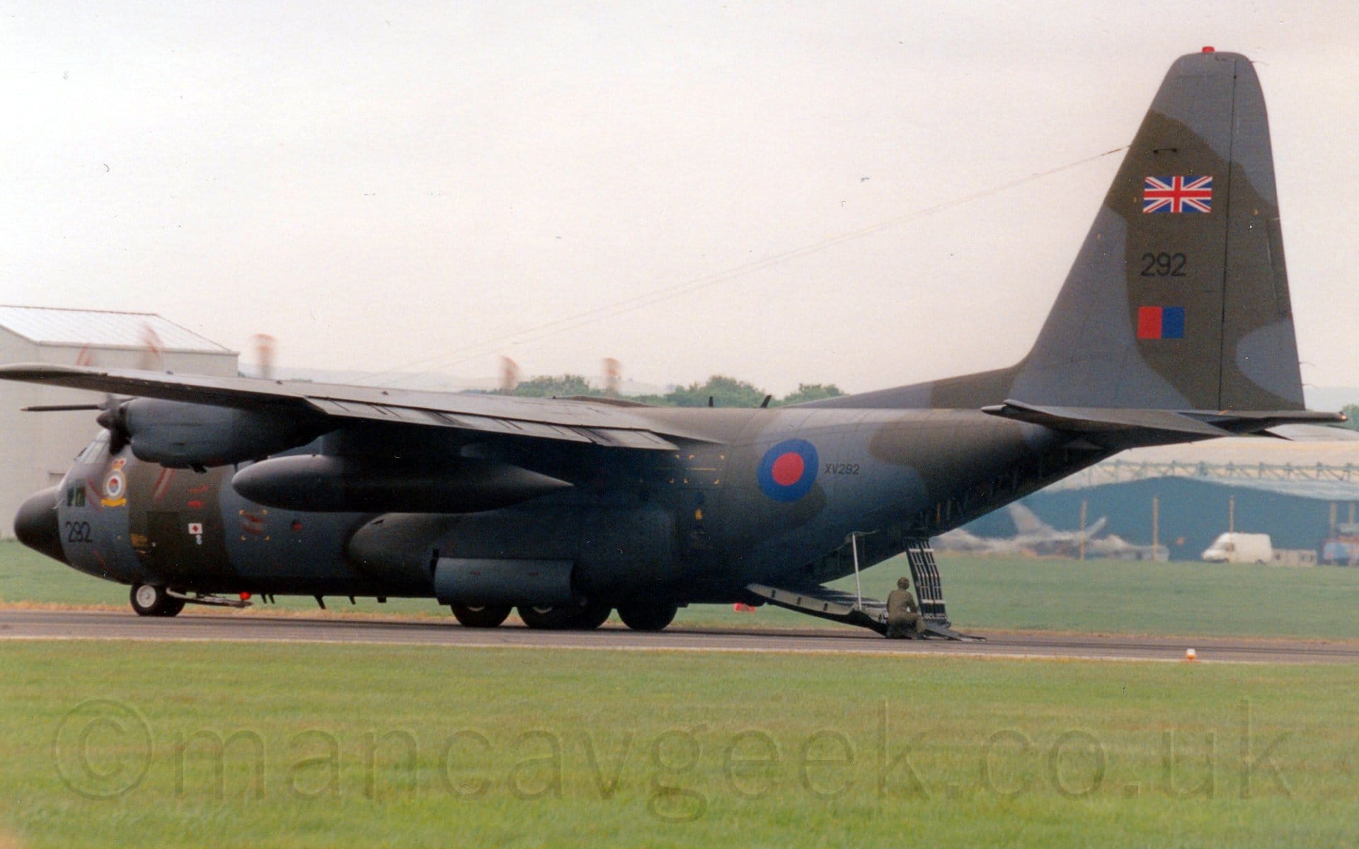 Side view of a high-winged, 4 propellor-engined military cargo plane parked on a black runway, with the red and white tips of the propellors a spinning blur indicating they are running full tilt, flaps extended from behind the wings, and the rear cargo loading ramp almost fully deployed, with a man wearing olive-green fatigues crouched by the end of the ramp. The plane is covered in an olive-green and grey camouflage scheme, with a blue and red roundel on the rear fuselage, next to the black serial number "VX232". There is a black number "232" on the nose under the cockpit, repeated on the tail, where it is sandwiched between a British flag above, and a red and blue fin flash below. Green grass fills the foreground, with more grass leading up to a tall grey hangar on the left, and a lower blue hangar on the left, with a couple of grey fighter jets in front of the latter. Grey sky fills the rest of the frame.