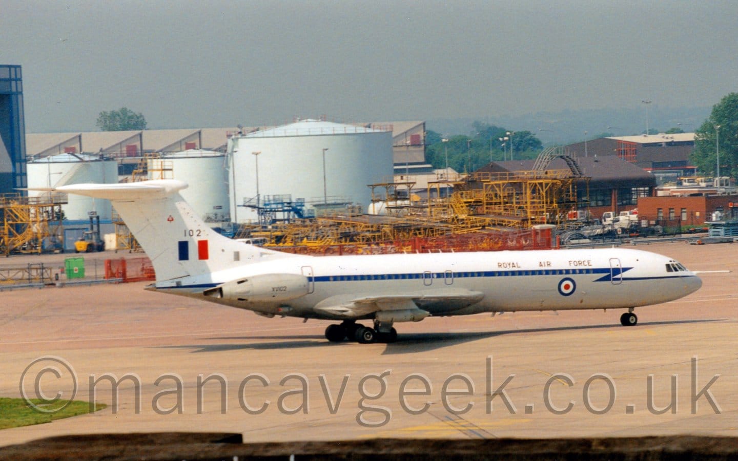 Side view of a 4 engined jet airliner taxiing from left to right. The plane's upper surfaces are white, while the lower sections are grey, with a dark blue stripe running along the body, covering the passenger cabin windows, separating the 2 colours. There are black "Royal Air Force" titles on the upper forward fuselage, above a large blue, white, and red roundel on the lower body. The white tail has the large black number "102" above a blue, white, and red fin flash. The plane's engines are mounted in pairs on the sides of the rear fuselage, slightly in front of the tail, and have the black serial "XV102" on them. Grey concrete apron fills the foreground, with random yellow metal frames in the background in front of pale blue fuel tanks and other buildings, under blue-grey sky.