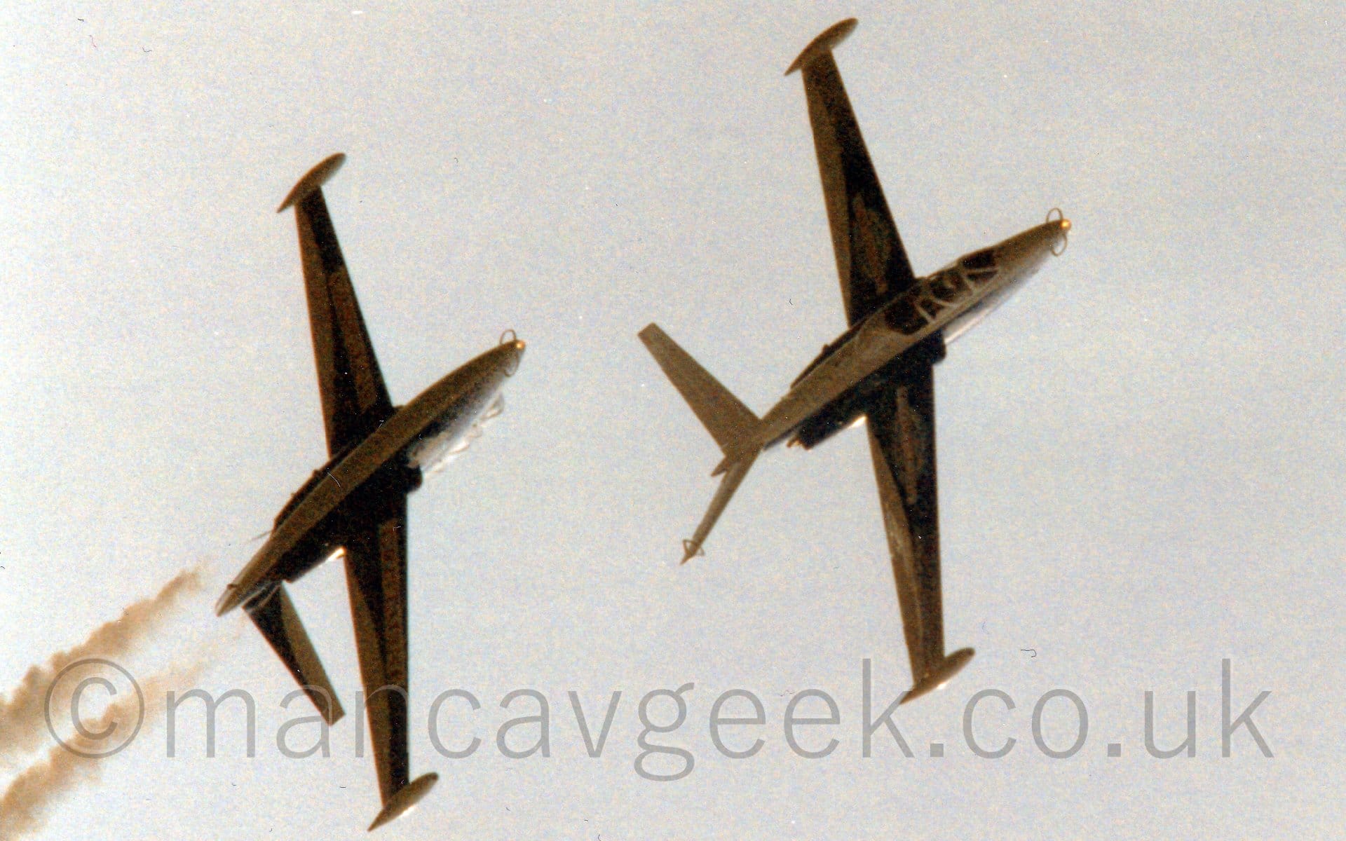 Slightly grainy view from below of a pair of jet trainer aircraft flying in formation from left to right, one above the other, and inverted. The planes are mostly dark blue, with white upper and lower surfaces and v-tail, with indistinct red and white logos on the body and wings. Grey-blue sky fills the rest of the frame.