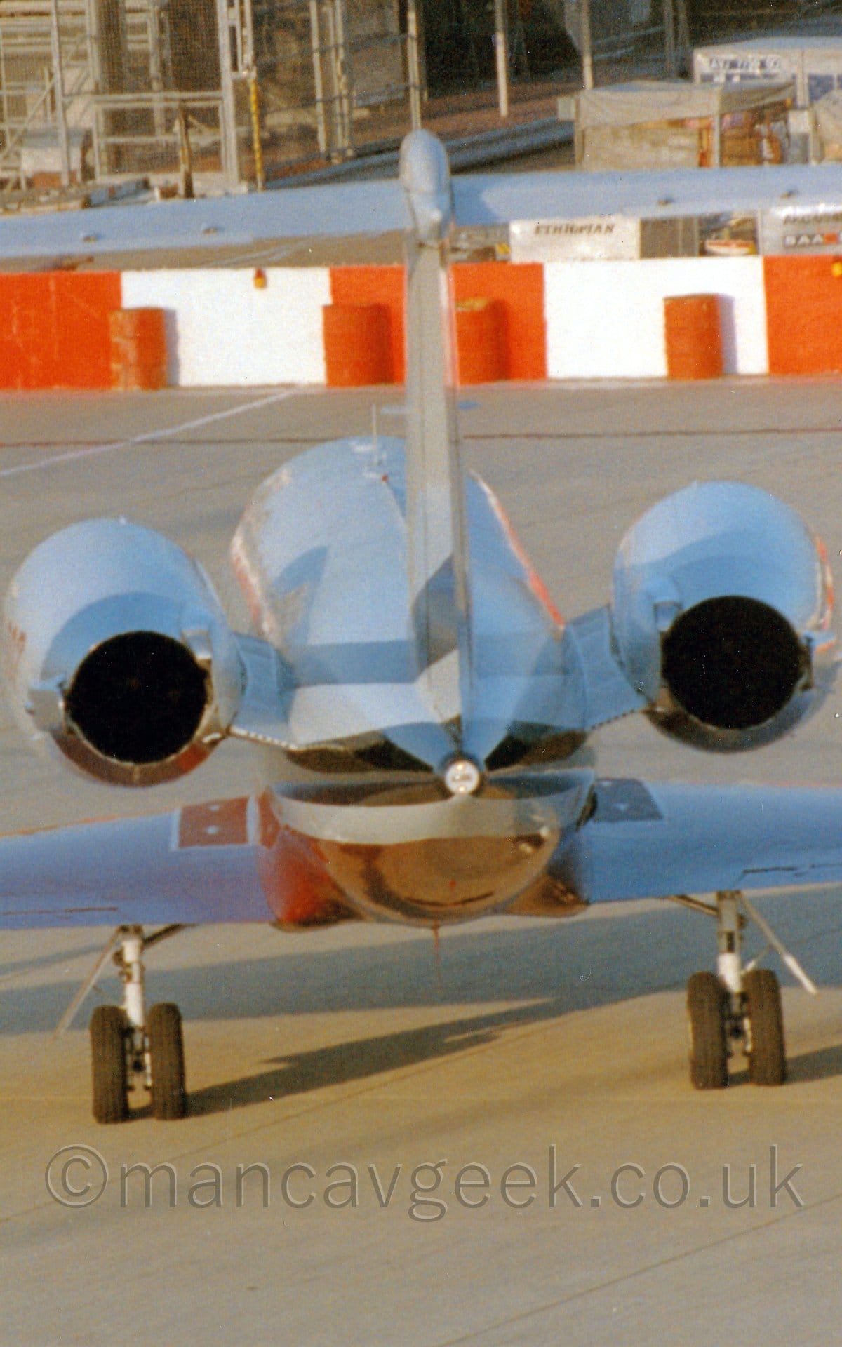 Rear shot of a twin engined BizJet parked on concrete apron facing away from the camera. The plane is mostly white, with red and black stripes running along the body, under the engines mounted on the sides of the rear fuselage. There are red and white barricades in the background, forming the perimeter of a building site.