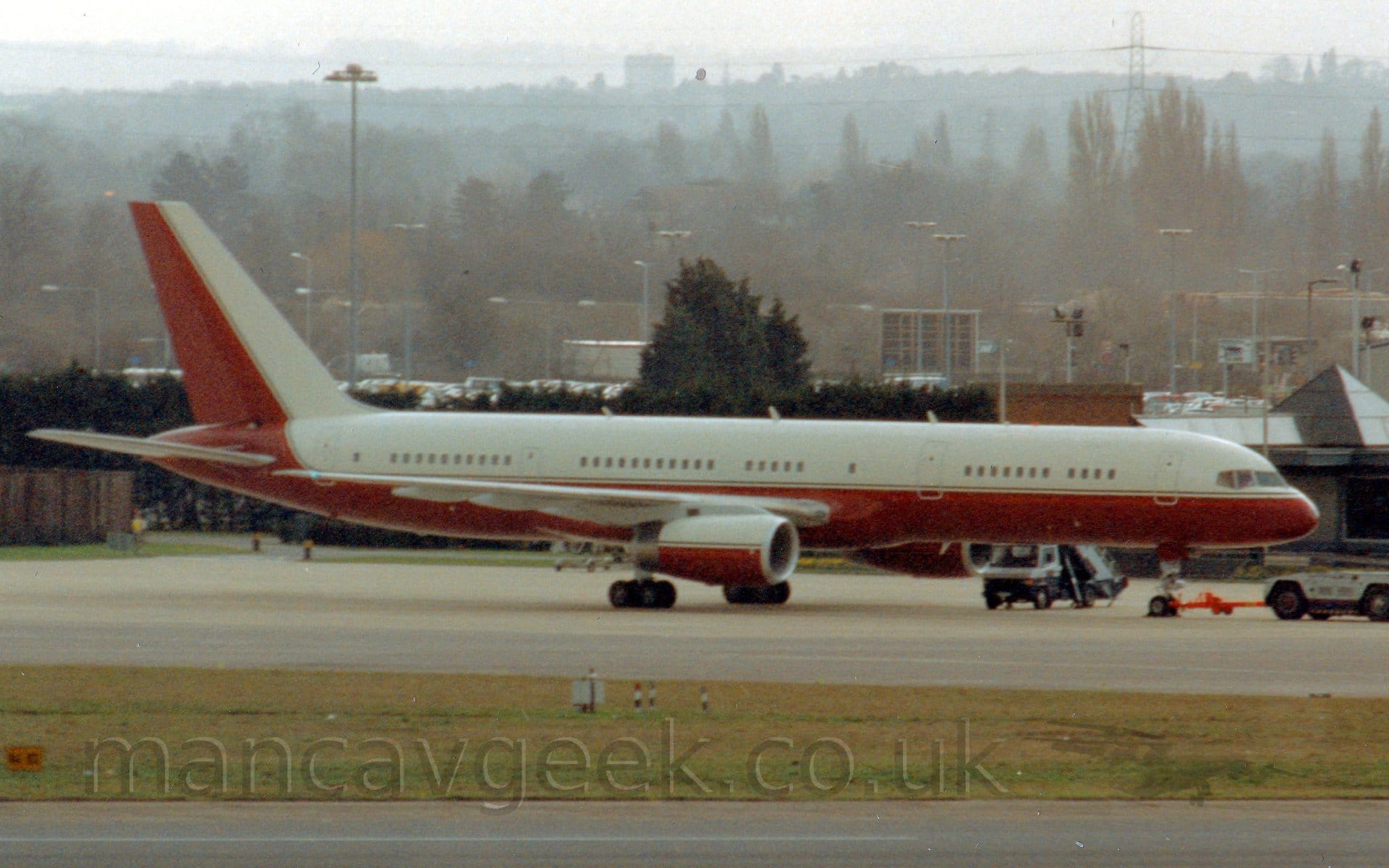 Side view of a twin engined jet airliner parked facing to the right. The plane has white upper surfaces and red lower surfaces, with the tail being white at the front and red at the rear. There is a grey and dark blue tug attached to the planes nose-wheel by a long red pole. Green grass stretches across the foreground, separating the runway from a taxiway, with trees and buildings stretching into the distance in the background, slowly vanishing into hazy skies.