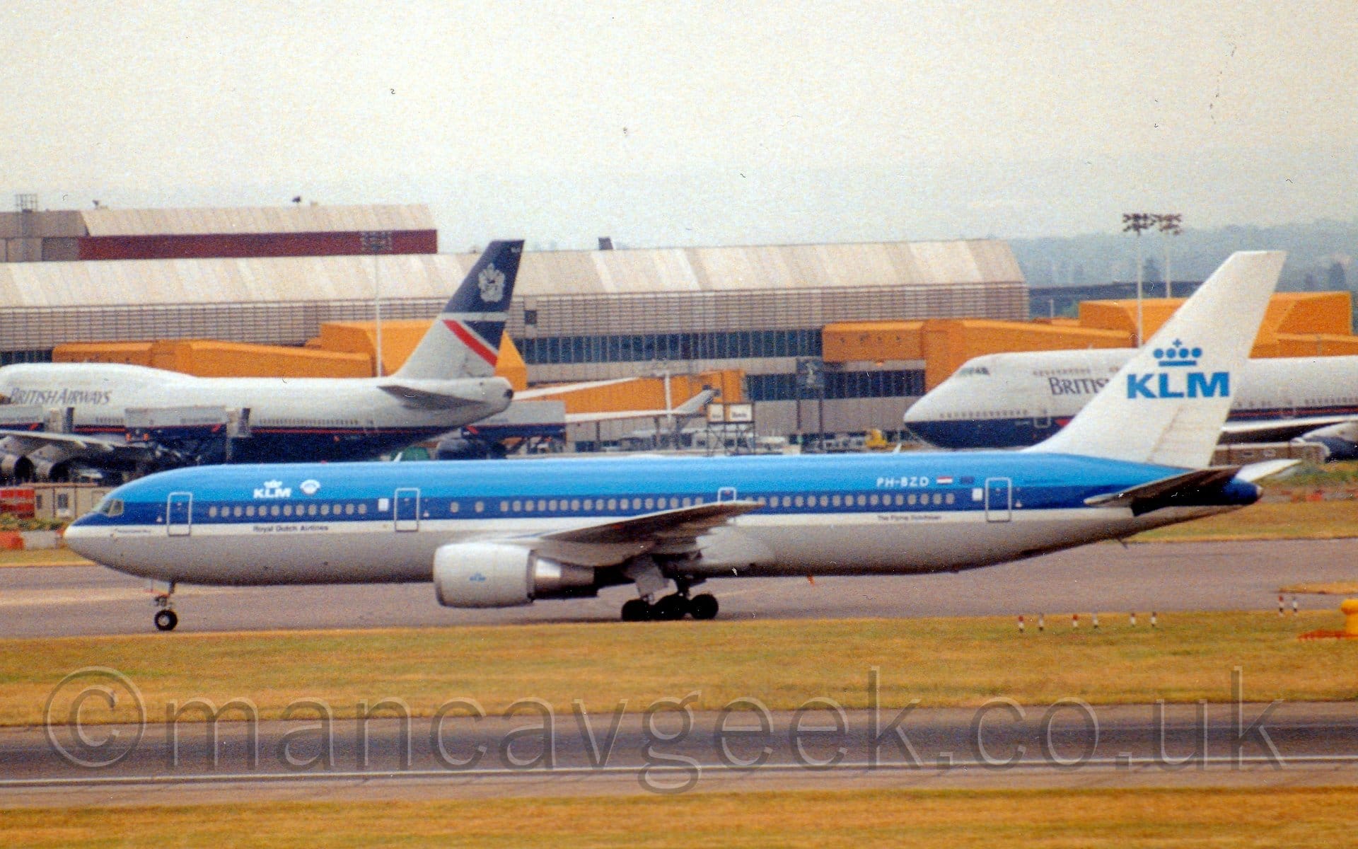 Side view of a twin engined jet airliner taxiing from right to left. The plane has blue upper surfaces, with a grey belly, separated by a white stripe. There are small, white "KLM" titles under a stylised white crown on the upper forward fuselage, repeated in blue in a larger form on the white tail. Slightly yellowing grass lines a black runway in the foreground, while a grey terminal building with large yellow airbridges hanging off the side stretches across most of the background from the left, with a couple of grey and dark blue jet airliners in front. Grey sky fills the rest of the frame.
