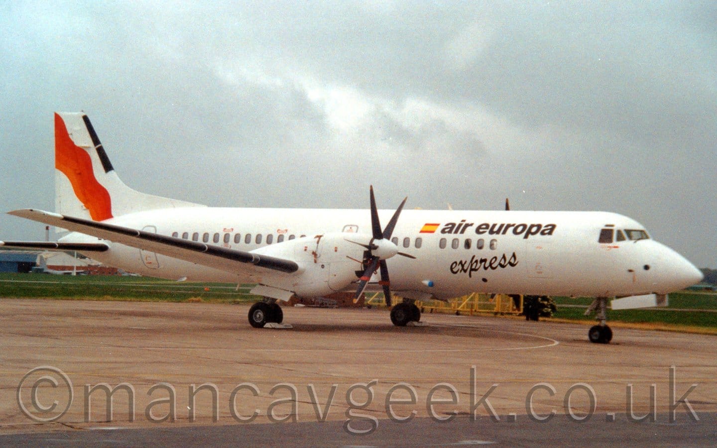 Side view of a twin propellor-engined parked on grey concrete apron facing to the right. The plane is almost entirely white, with black "Air Europa" titles on the upper forward fuselage next to a small Spanish flag, with black, seemingly hand-written "Express" text below. An orange and red wavy diagonal stripe is on the white tail. Grey concrete apron fills the foreground, with grass in the background visible under the plane, with cloudy grey sky above.