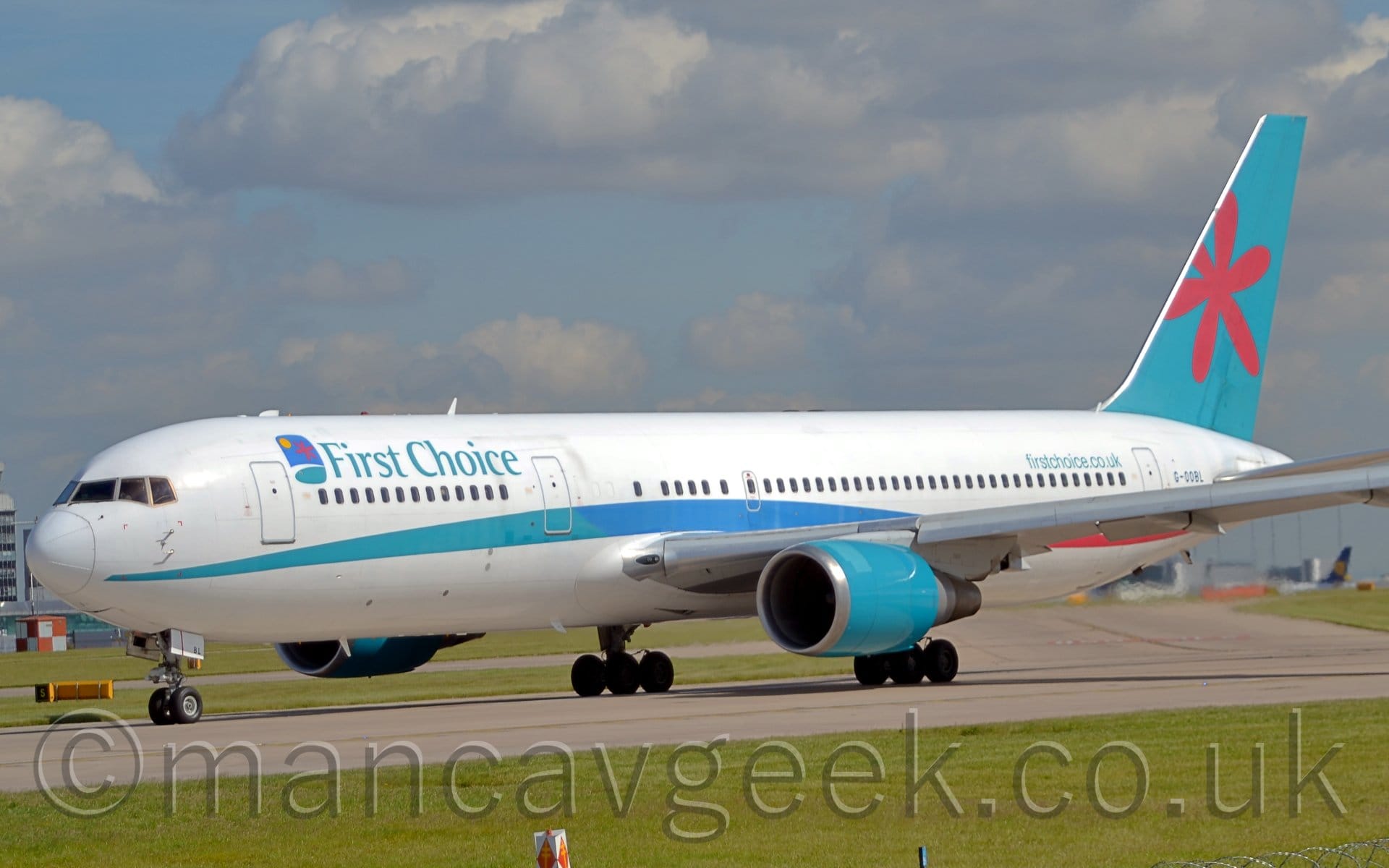 Side view of a twin engined jet airliner taxiing from right to left along a grey runway. The plane is mostly white, with a multi-tone green/blue stripe running along the body, and green "First Choice" titles on the upper forward fuselage. There is additional green text "FirstChoice.co.uk" on the upper rear fuselage, and black registration "G-OOBL" just aft of the rear door. The tail is a green/blue, with a stylised red flower in the middle. The engine pods under the wing are the same green blue. Lush green grass lines the runway in the foreground, with pale blue sky and fluffy clouds filling most of the background.