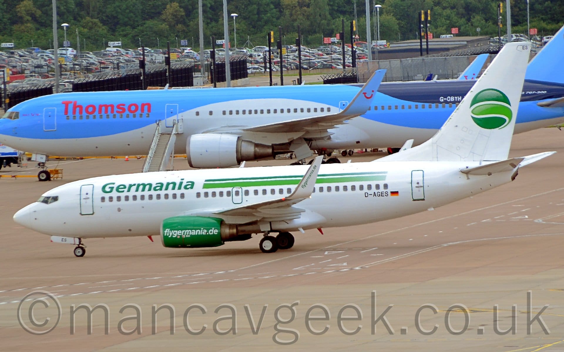 The white tail has a dark green circle with a lighter green slice in the middle. The black registration "D-AGES" is on the lower rear fuselage next to a small German flag. The engine pods slung under the wings are bright green, with white "FlyGermania.de" text. The white upturned wingtips have that same text, but this time in green. Grey concrete apron fills the foreground, with a larger, blue and white twin engined airliner stretching across the frame in the background, with a sprawling ground-level car park in front of trees in the distance.