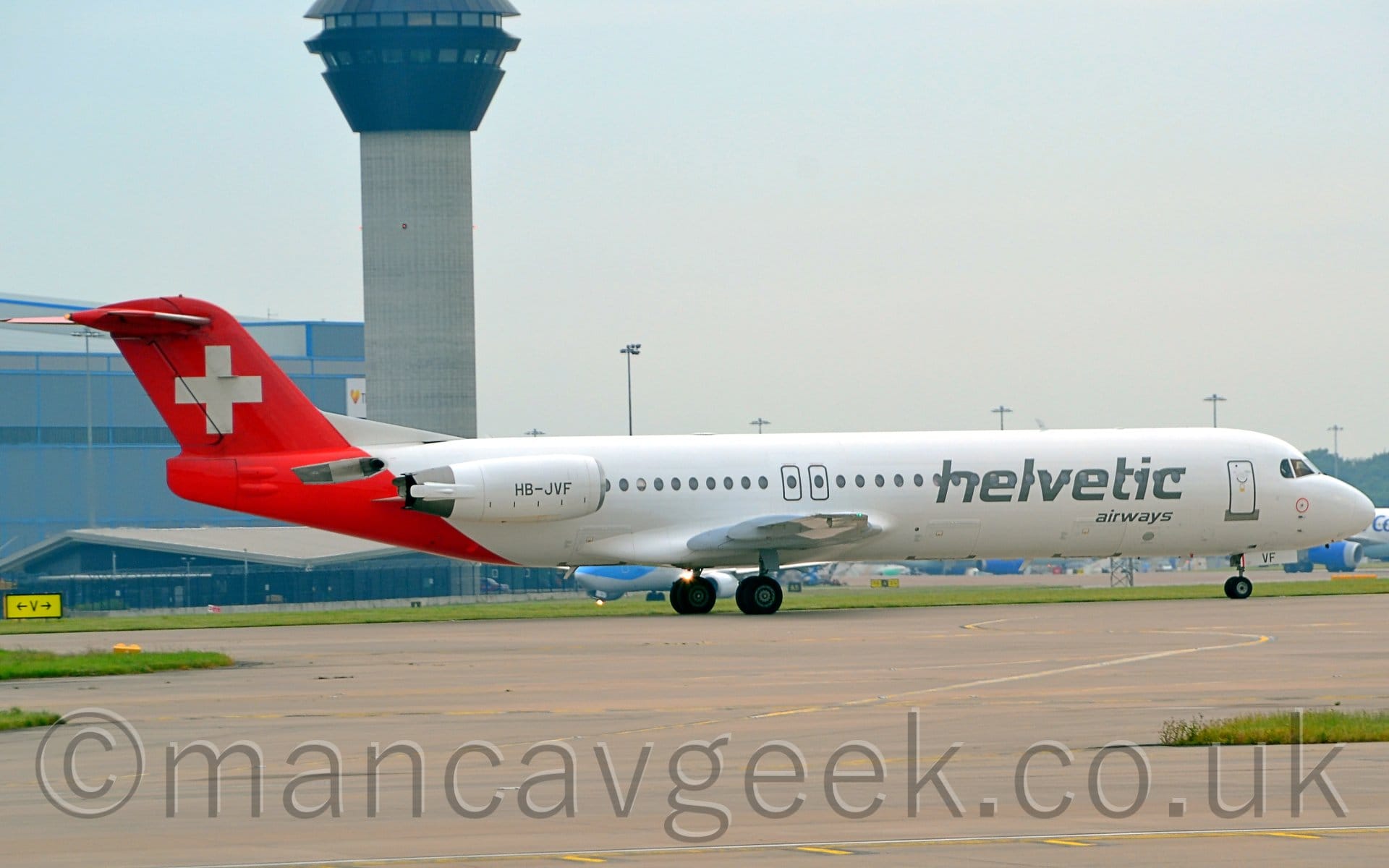 Side view of a twin engined jet airliner taxiing from left to right. The plane is mostly white, with large grey "Helvetic" titles covering the forward passenger cabin windows, above smaller grey "Airways" text. The registration "HB-JVF" is on the white engine pods, mounted on the sides of the rear fuselage. The rear fuselage and tail are red, with a white cross in the middle.