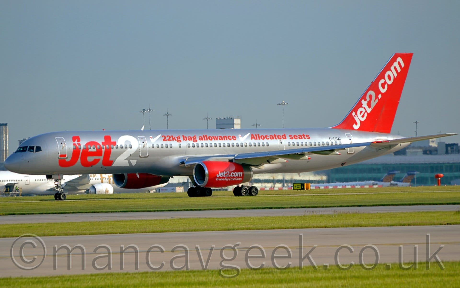 Side view of a twin engined jet airliner moving from right to left. The plane is mostly a shiny silvery-grey, with red "jet2" billboard-stye titles (the "2" in white) on the forward fuselage, with additional red "22kg bag allowance" and "Allocated Seats" text next to large white check marks on the upper fuselage. There are large white and grey "Jet2[dot]com" titles on the bright red tail, as well as on the engine pods mounted under the wings. The engines also have smaller, white "Friendly low fares" in an almost hand-written font. Taxiways lined with vivid green grass stretch across the foreground, while a grey terminal building stretches across the background, with several other planes visible parked in front, all under blue-grey sky.