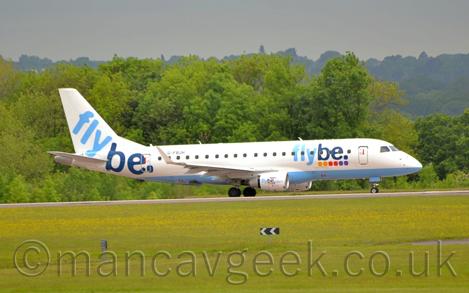 Side view of a twin engined jet airliner taxiing from left to right along a grey runway. The plane is mostly white, with a pale blue belly, and 2-tone, dark and light blue "FlyBe" titles above some coloured dots on the forward fuselage, with larger "FlyBe" titles on the rear fuselage and tail. There are additional "FlyBe[dot]com" titles on the engine pods mounted under the wings. An expanse of green grass fills the foreground, with green trees stretching across the background, under grim grey skies.
