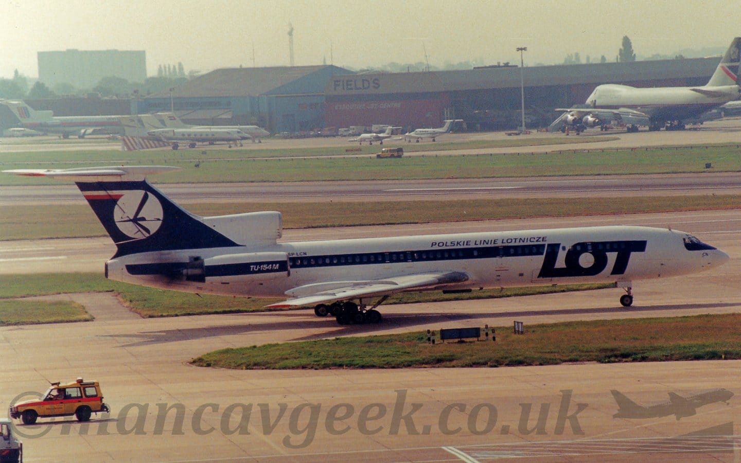 Side view of a 3 engined jet airliner with the engines mounted on the rear fuselage, taxiing from left to right. The plane is mostly white, with a thick blue stripe running along the body, covering the passenger cabin windows, as well as over the engines mounted on the sides of the fuselage. There are also large, blue "LOT" titles on the forward fuselage, with smaller blue Polish titles that translate into English as "Polish Air Lines". The tail is the same dark blue, with a white circle containing a stylised image of a blue flying bird. Grey concrete apron fills the foreground, with green grass bordering taxiways and a runway in the background leading up to a red and grey hangar next to a blue and grey hangar, both with planes parked in front, under bright but hazy sky.