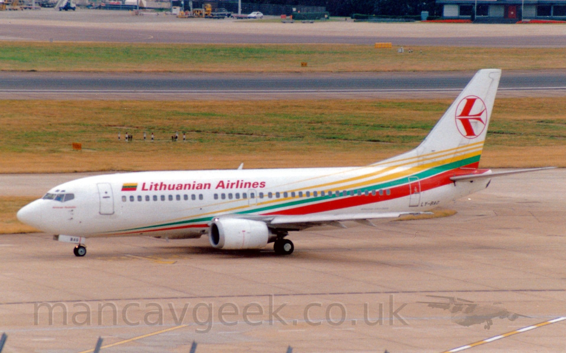 Side view of a twin engined jet airliner taxiing from right to left. The plane is mostly white, with yellow, green, and red diagonal stripes running from the lower forward fuselage to the upper rear fuselage and up into the lower reaches of the tail. There are red "Lithuanian Airlines" on the upper forward fuselage, next to a small Lithuanian flag. The black registration "LY-BAG" is on the lower rear fuselage. The white tail has the red outline of a circle containing a stylised red flying bird. Grey concrete apron fills the foreground, with wide swathes of grass either side of a black runway in the background.