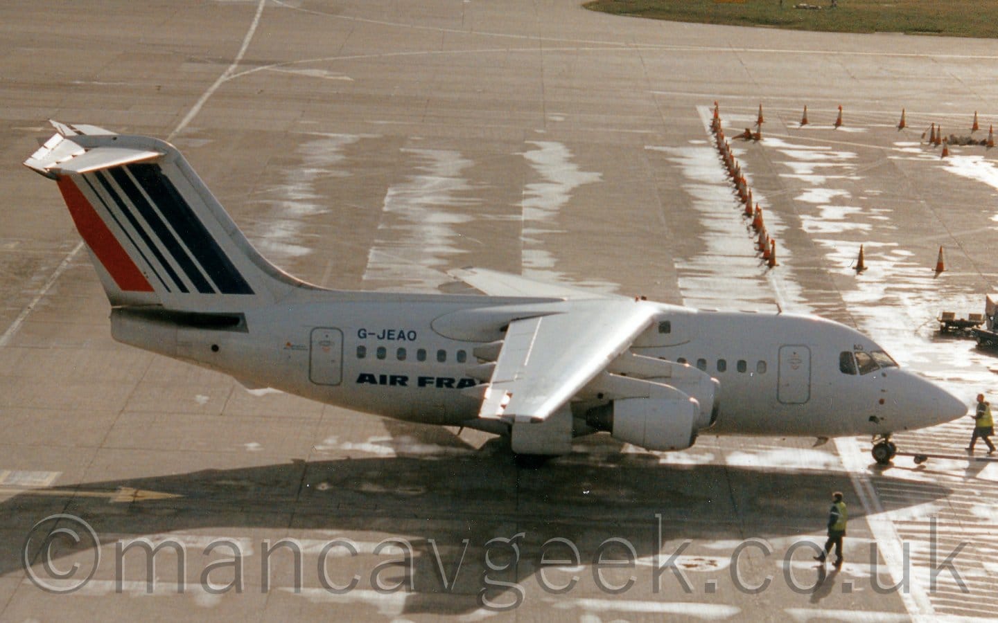 Side view of a high-winged, 4 engined jet airliner facing to the  right but being pushed back to the left by an out-of-frame tug attached to the nose-wheel by a long pole. The plane is mostly white, with dark blue "Air France" titles on the lower rear fuselage, with the black registration "G-JEAO" on the upper rear. The tail has a series of dark blue diagonal stripes, thicker towards the front, with a single red stripe at the rear. Grey concrete apron fills most of the rest of the frame, with a row of orange and white traffic cones in the background on the right, and a couple of people wearing hi-viz jackets wandering around. Bright patches on the ground suggest that, while the sun bay be shining brightly, as suggested by the strong shadow this plane casts on the ground, but it was raining not too long ago.