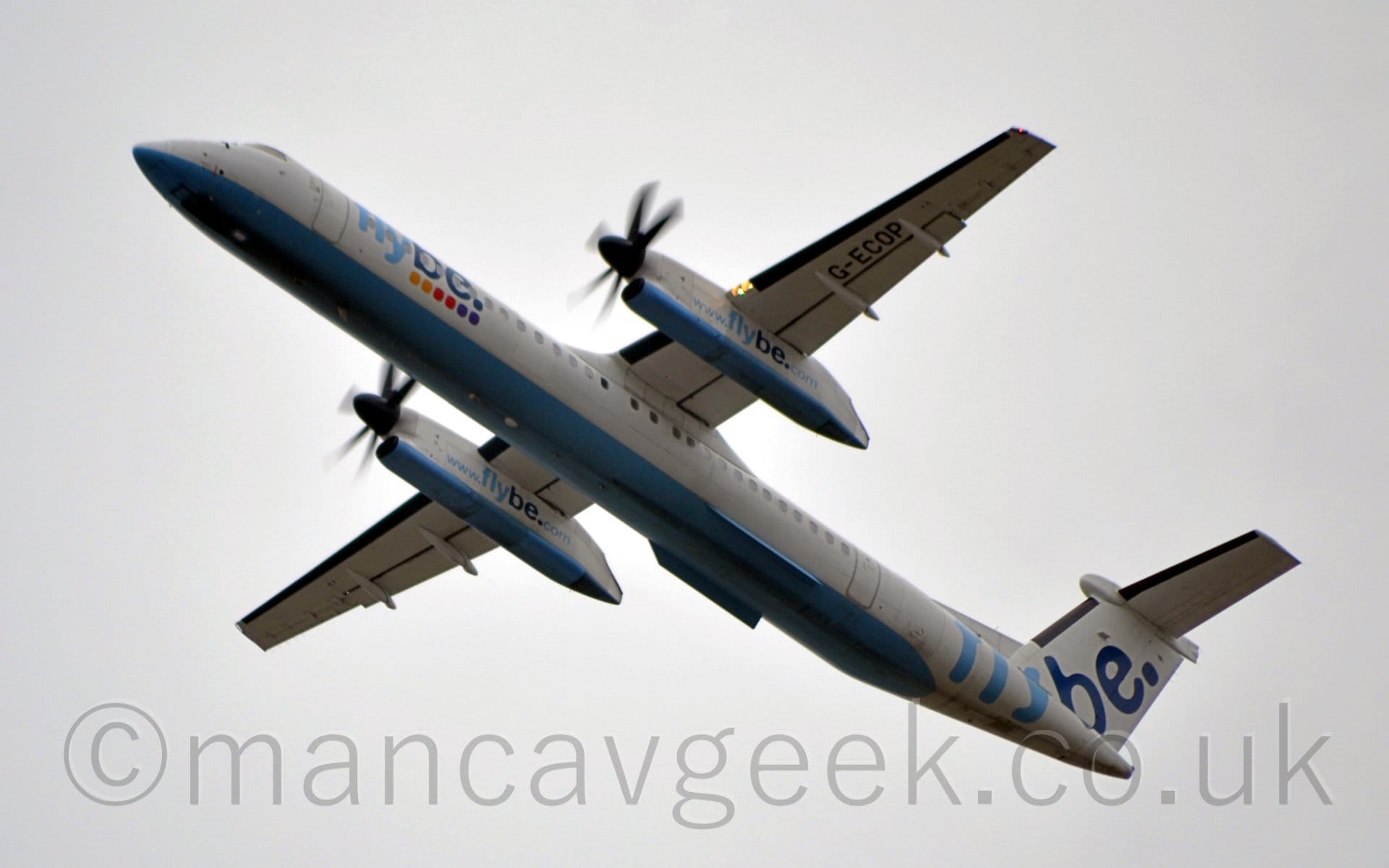 Low side view of a high-winged, twin propellor-engined airliner flying from right to left at a very low altitude, with flaps extended from the rear of the wings, the undercarriage already stowed away, and the nose raised sharply, suggesting it has just taken off. The plane is mostly white, with a pale blue belly, and light and dark blue "FlyBe" titles on the forward fuselage over a group of 6 coloured dot - 2 each yellow, red, and purple. There are larger "FlyBe" titles on the tail, as well as the website address "www.flybe.com" on the sides of the engine pods under the wings, which also have a pale blue lower section. The black registration "G-ECOP" is on the outer panels of the underside of the left wing. Cloudy grey sky fills the rest of the frame.