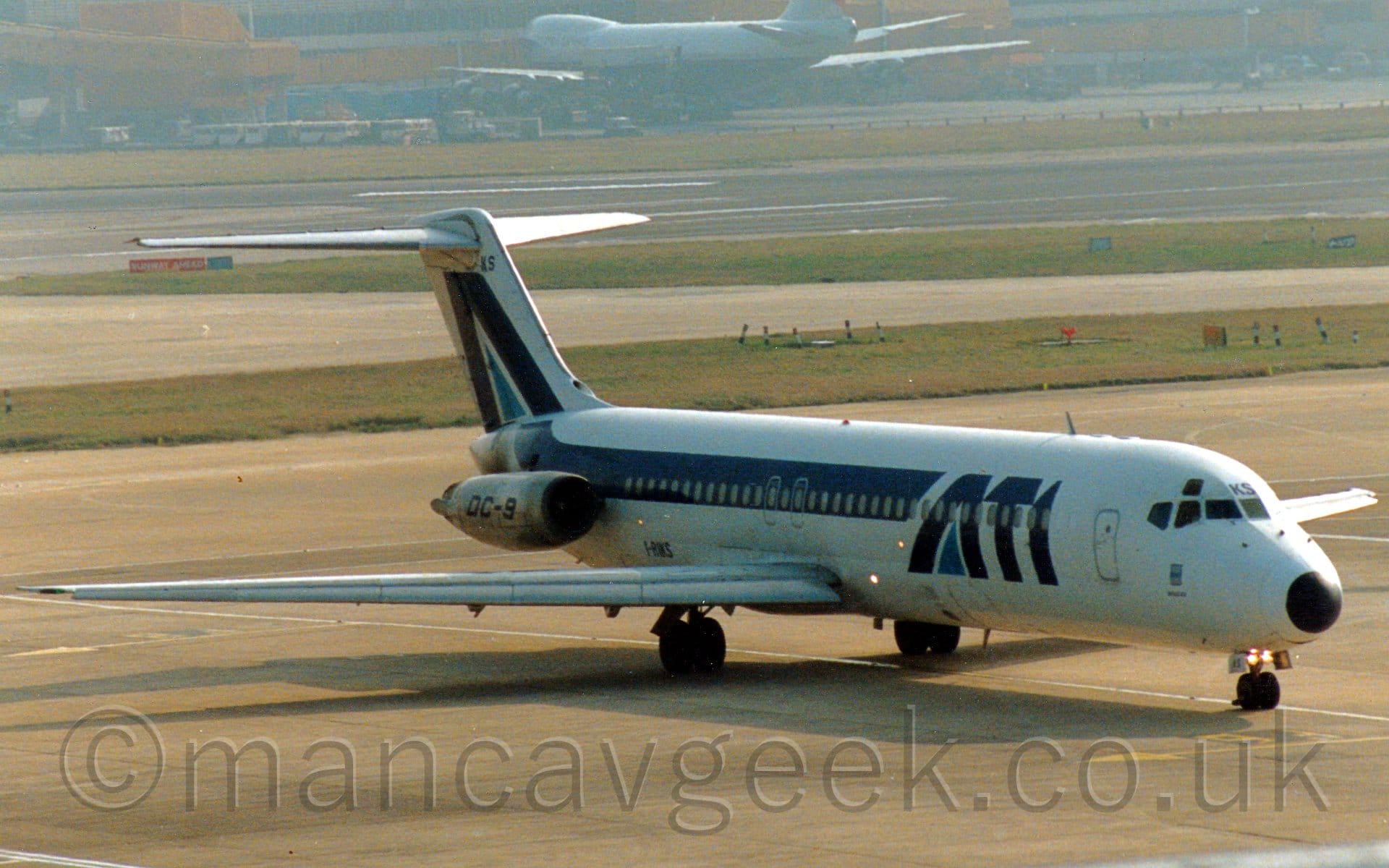 Side view of a twin engined jet airliner with the engines mounted on the sides of the rear fuselage, taxiing from left to right, following a white line on the taxiway. The plane is mostly white, with dark blue "ATI" titles on the forward fuselage, with a lighter blue triangle filling the lower part of the letter "A", and a dark blue stripe running along the body, turning up into the tail where it repeats that letter "A". There are black "DC-9" titles on the bare-metal engine pods on the sides of the rear fuselage. Grey concrete apron fills the foreground, with large areas of green grass lining taxiways and a black runway in the background, with a grey and yellow terminal building stretching across the frame in the distance, with a very large, grey and blue jet airliner parked nose in in front of it.