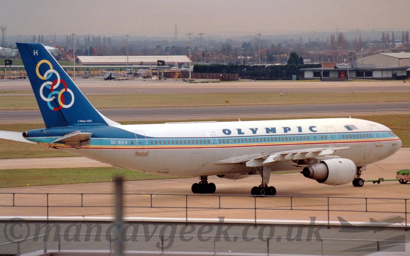Side view of a twin engined jet airliner facing to the right but being pushed back to the left by an almost out-of-frame green tug. The plane has white upper surfaces and a grey belly, with a light blue stripe running along the body covering the passenger cabin windows, with additional blue pinstripe above, and yellow and red pinstripes below. There are dark blue "Olympic" titles on the upper fuselage over the wings, with a stylised Greek flag partially covered by an EU flag between the forward and mid-cabin doors. The tail is dark blue, with a series of linked, multi-coloured rings, and the white letter "H" at the top. The grey flat roof of a building fills the bottom of the photo, while green grass lines a runway and a taxiway in the background, with trees and buildings leading off in to the distance, slowly vanishing into grey haze.