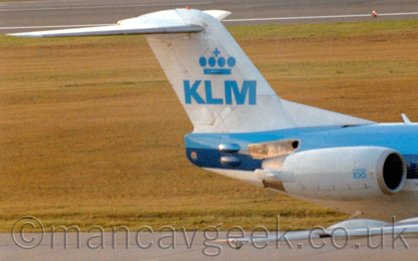 Close-up of the white tail of a twin engined blue and white jet airliner taxiing from left to right. The planes white engines are mounted on the sides of the rear fuselage, and have small "Fokker F100" text at the front. The planes tail is white, with large blue letters "KLM" under a stylised blue crown in the middle. Yellowing grass fills the background, with a black taxiway stretching across the top of the frame.