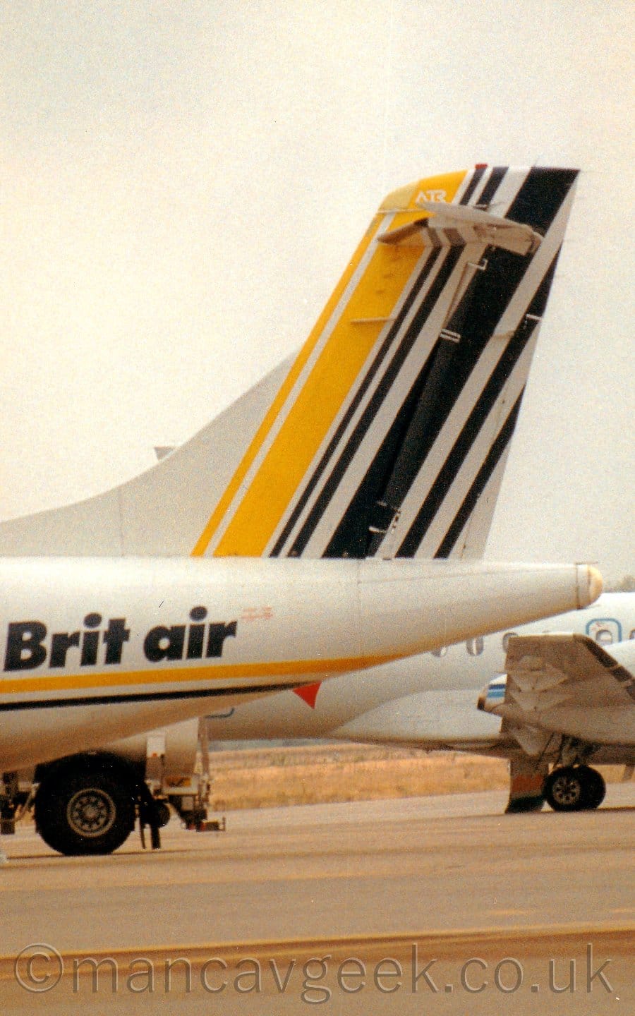 Close-up of the tail and rear fuselage of an airliner parked facing to the left. The plane is mostly white, with a thin blue and yellow stripe running along the body, with blue "Brit Air" titles on the rear fuselage, and yellow and blue diagonal stripes on the tail. There is another white plane parked behind, with cloudy white sky filling the rest of the frame.