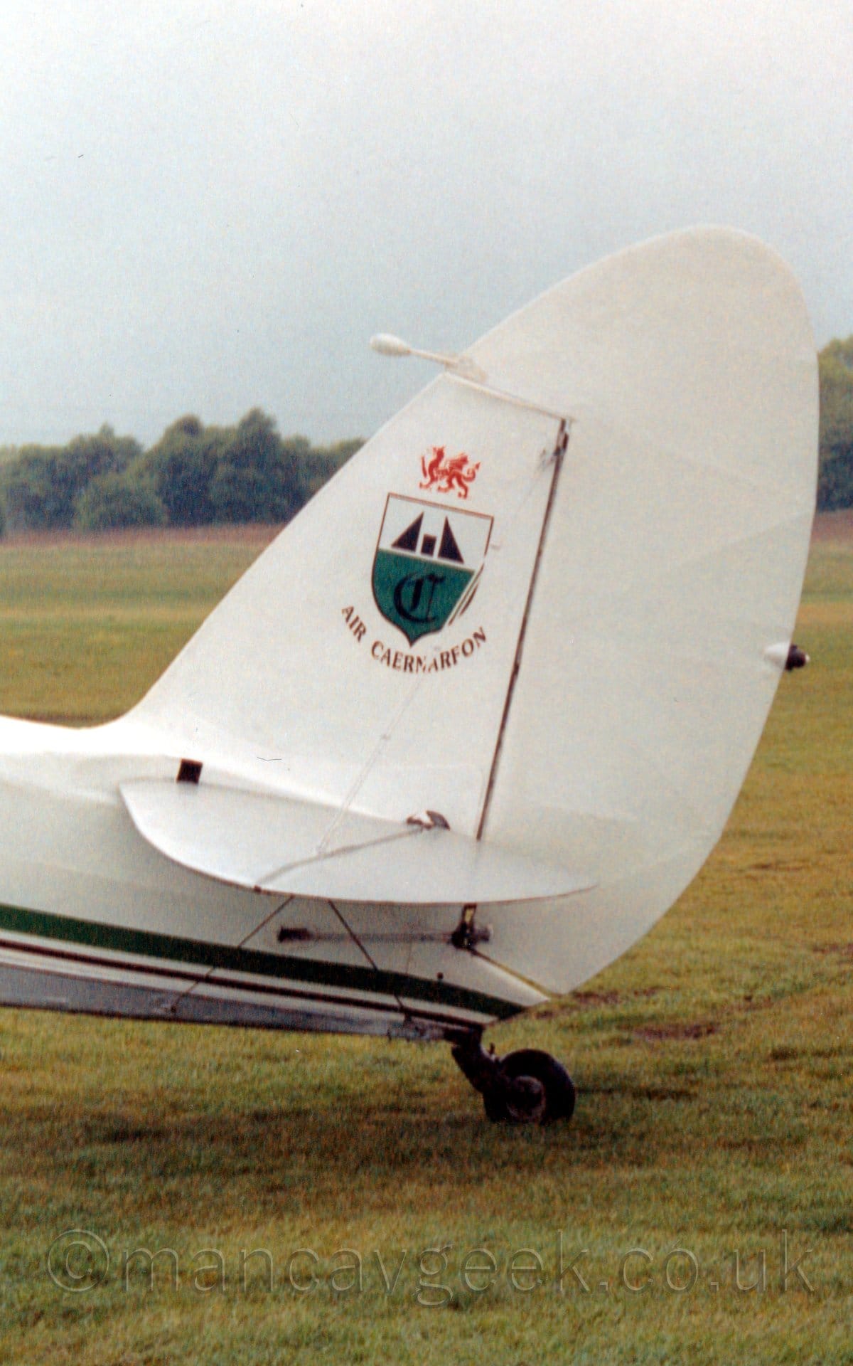 Close-up of the tail and rear fuselage of an airliner parked facing to the left. The plane is mostly white, with a thin blue and yellow stripe running along the body, with blue "Brit Air" titles on the rear fuselage, and yellow and blue diagonal stripes on the tail. There is another white plane parked behind, with cloudy white sky filling the rest of the frame.