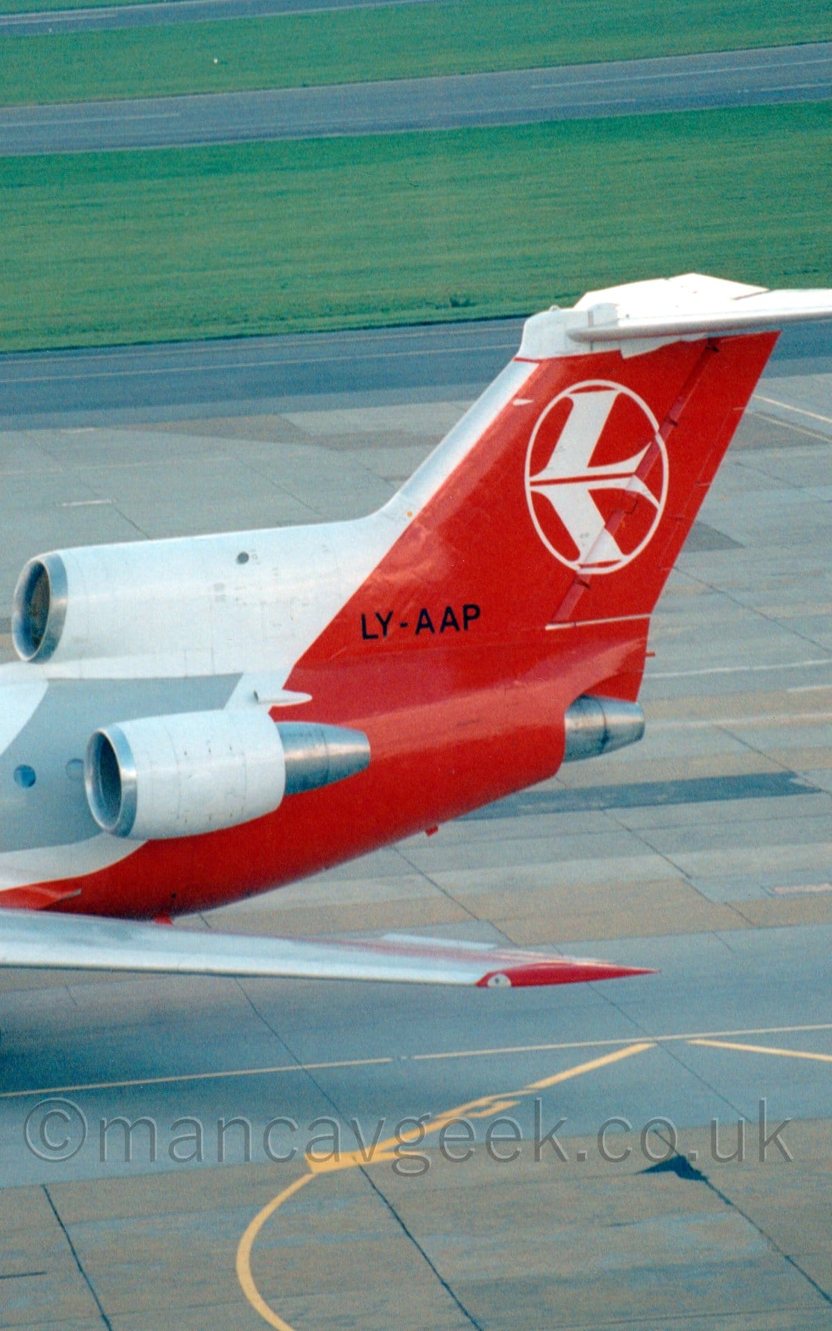 Close-up of the rear fuselage and tail of a 3 engined jet airliner taxiing from right to left. The plane has a white body with a red belly and a grey stripe covering the passenger cabin windows. The tail and rear fuselage are red, with a stylised white bird in the white outline of a circle in the middle of the tail. The white engines are mounted on the top and sides of the rear fuselage , with the centre engine intake being built in to the front of the tail, but exhausting below it. The black registration "LY-AAP" is at the bottom of he tail. Grey and black concrete apron fills the foreground, with black taxiways divided by large grassed areas fills the background.