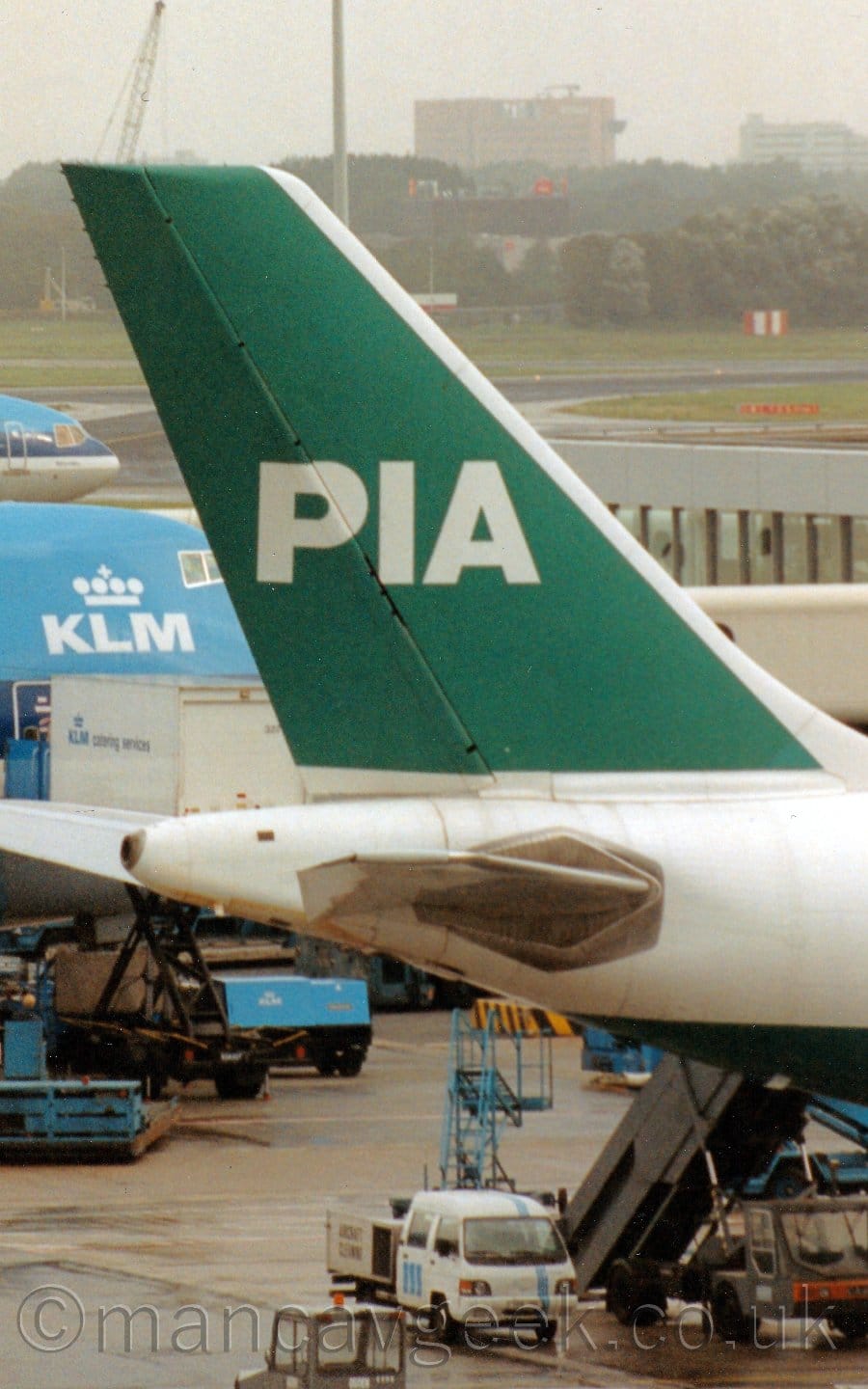 Close-up of the green tail of a twin engined jet airliner parked on it's stand facing to the right. The plane has a white body with a green body, while the tail is green with a white "PIA" text logo. A grey terminal building is in the background, with a couple of blue planes parked against it.