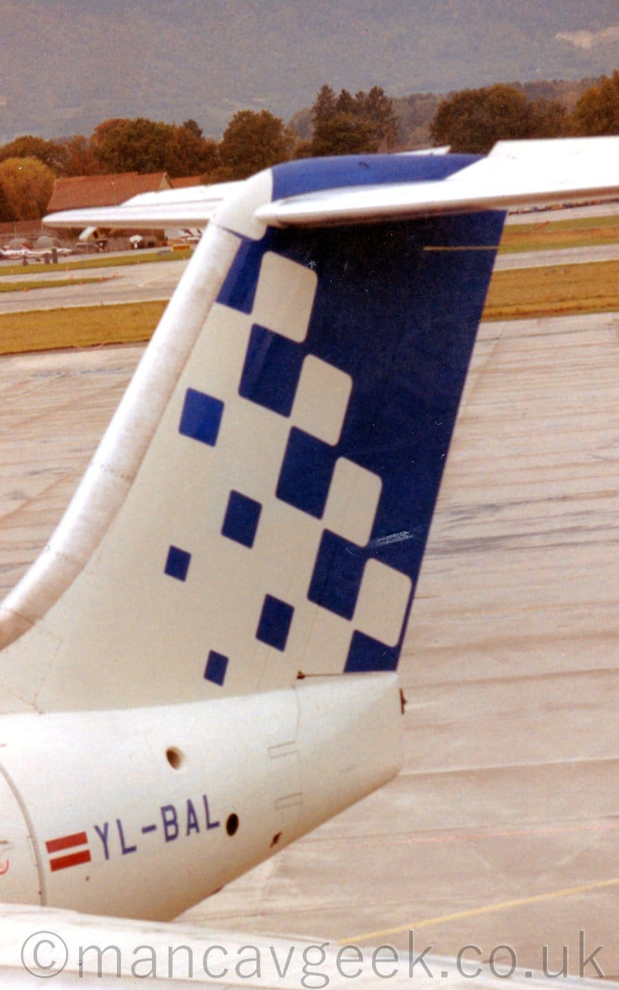 Close-up of the white tail and rear fuselage of a jet airliner parked facing to the left. Small blue squares at the front of the tail slowly grow bigger as they move rearwards, until they fill the whole of the tail. The blue registration "YL-BAL" on the rear fuselage, next to a Lithuanian flag. Grey concrete apron in the background leads off into the distance, with mountains beyond.