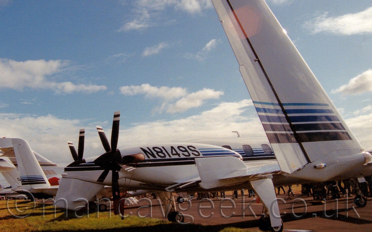 Close-up of the twin tails of a twin engined BizProp parked on balck tarmac facing to the right. The plane is mostly white, with a series of light and dark blue stripes running along the body and over the engines, which are mounted on the rear of the wings, facing backwards. The black registration "N8149S" is painted on the sides of the engine cowlings. As well as the twin tails on the ends of the wings, there is a small ventral tail under the rear fuselage - whether that is for additional stability, or to stop the propellor blades hitting the ground on take off or landing is unknown, at least to me. (It's probably a bit of both!) The tails of several other planes can be seen in the background on the left, and the feet of people in the crowd can be seen under this plane. Gorgeous blue sky with scattered fluffs of cloud fill the rest of the frame.