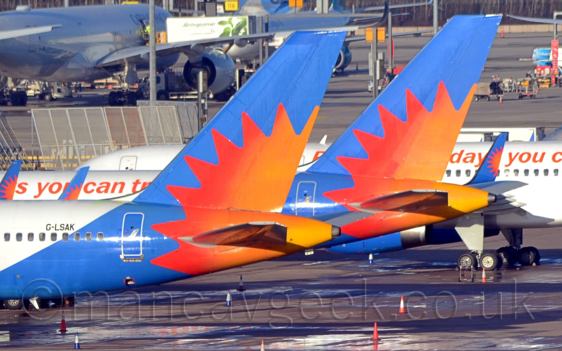 Close-up of the rear fuselage and tail of a pair of near-identical jet airliners parked facing to the left. The planes have white bodies, with a blue rear fuselage and tail, with a large red and orange sun burst. The nearest plane has the black registration "G-LSAK" on the upper rear fuselage. The second plane has some red, fairly indistinct text running along the body. There is a third plane from the same airline showing most of it's fuselage behind them. Another white jet airliner is in the background on the left. Bright sunlight floods the frame, causing reflections all over the place.