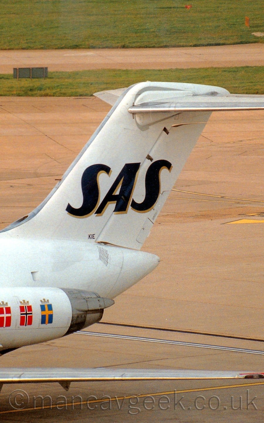 Close-up of the white rear fuselage and tail of a twin engined jet airliner with the engines mounted on the sides of the rear fuselage. The plane is parked facing to the bottom left of the frame. There are 3 flags on the engine pods, on the left is a red flag with a white cross, in the middle is a red flag with a black cross that is outlined in white, and on the right is a blue flag with a yellow cross. Each flag has a gold crown on the top. The tail is white, with large black letters "SAS" with a gold drop-shadow, with very small letters "KIE" at the base of the tail. Grey concrete apron fills the background, with a large grass area at the top of the frame.