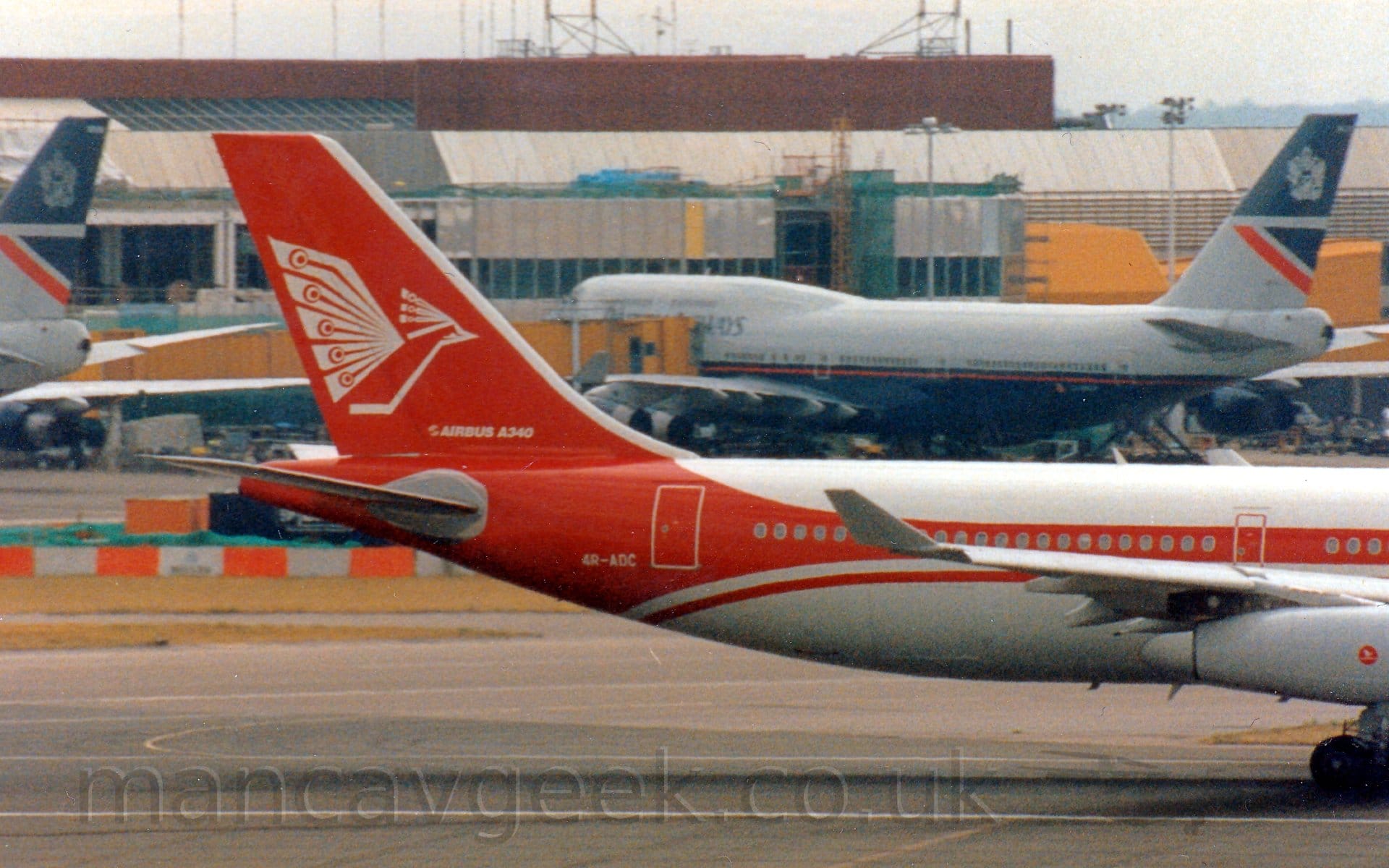 Side view of the rear fuselage and tail of a 4 engined jet airliner moving from right to left at a high speed. The plane is mostly white, with a thick red stripe running along the body, sweeping up into the tail and around the lower rear fuselage. There is a stylised white flying bird on the tail, above white "Airbus A340" titles, and the white registration "4R-ADC" on the lower rear fuselage. In the background, a grey terminal building stretches across the frame, with bright yellow airbridges attached to a very large, grey and dark blue jet airliner, with the tail of a similar plane coming in to view on the left of the frame.