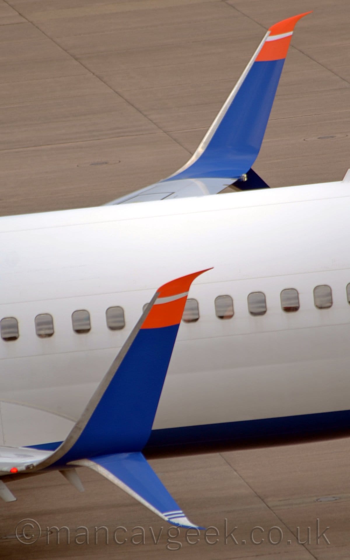 Close-up of the up-turned wing-tip of a twin engined jet airliner taxiing from right to left. The plane is white, with a blue belly. The tips of the wings curve upwards, painted the same blue, with the top section painted a  bright red with a white stripe. A smaller section also curves downwards, with white stripes at the tip. Grey concrete apron fills the rest of the frame.