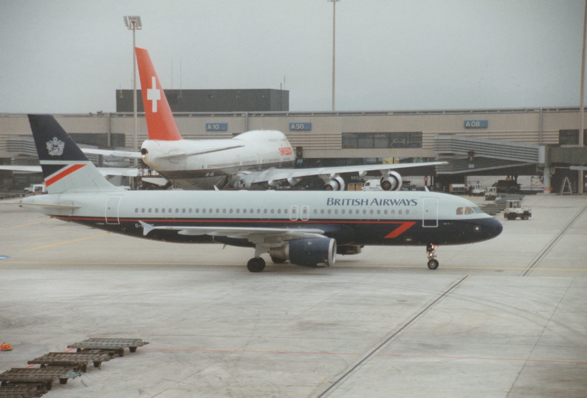 Side view of a twin engined jet airliner, taxiing from left to right, following a yellow line on the concrete apron. The plane has grey upper surfaces, with a dark blue belly with a red pinstripe, and dark blue "British Airways" on the upper fuselage. The colours reverse on the tail, with the upper half being dark blue, and grey at the bottom, with a dark blue and red triangle in the middle. Grey concrete apron fills the foreground, , with a sandy brown and grey terminal building stretching across the background, with a very large, white and brown 4 engined jet airliner with a red and white tail parked facing that building. Grey sky fills the rest of the frame.